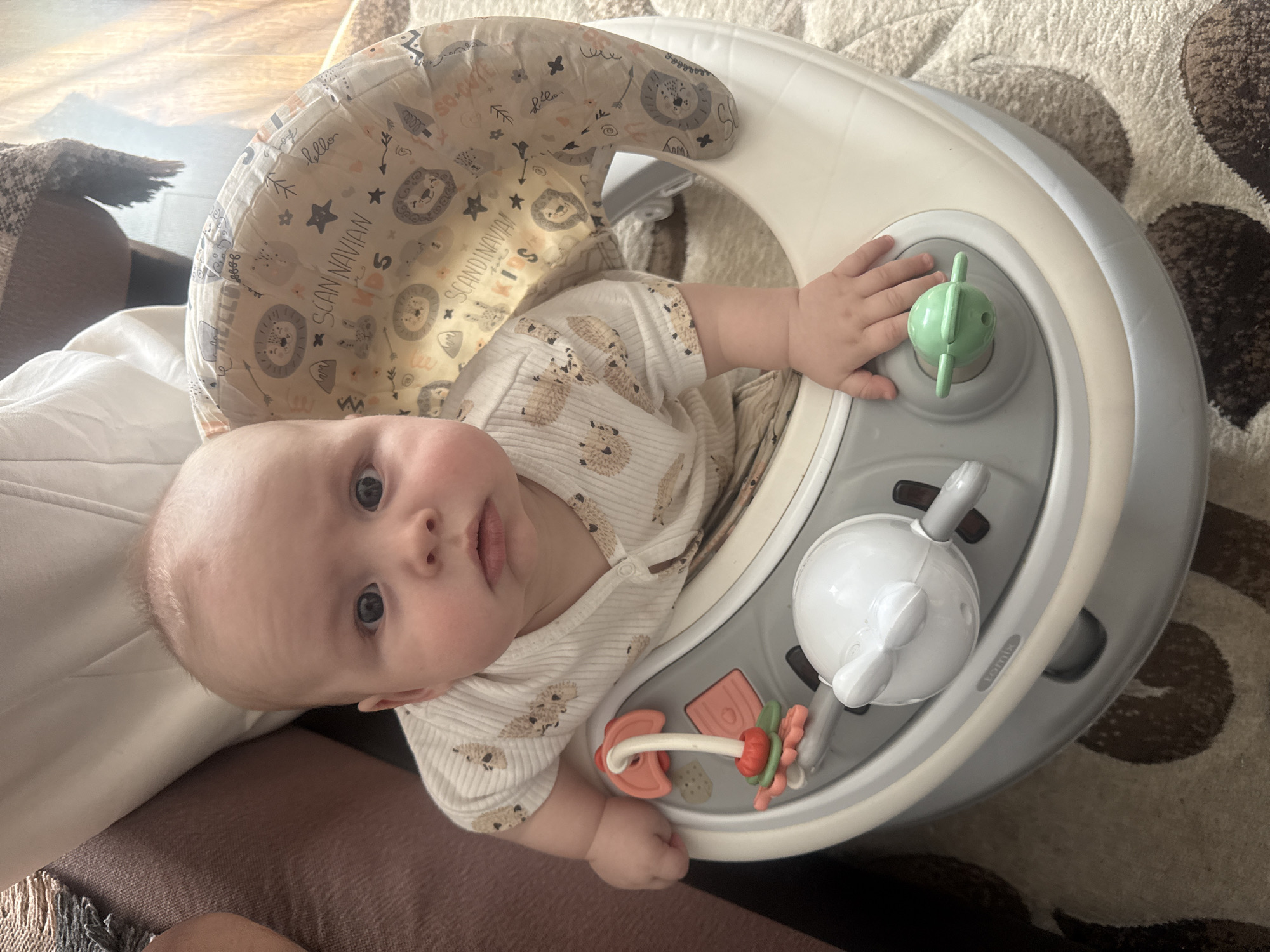 An overhead shot captures a baby seated in a light gray and white activity center indoors. The baby, an infant or young toddler with light skin, blue eyes, and light hair, looks directly at the camera with an alert, neutral expression. They are dressed in a light-colored, ribbed long-sleeved onesie adorned with a pattern of small brown hedgehog-like creatures.
The activity center features a fabric seat with a light-toned background patterned with various illustrations including lions, trees, stars, hearts, and arrows. Visible text on the fabric includes "SCAN NAVIAN," "KIDS," "hello," and "SCANDINAVIAN." The activity tray holds several toys: a light green propeller-like spinner, a white spherical toy with gray accents, and a red/orange and green teether/rattle. A small pink rectangular block is also seen on the tray. The baby's left hand rests near the green spinner, while the right hand is close to the teether. A faint "tomix" label is present on the tray.
The setting appears to be a cozy room, possibly a living room or nursery, with a soft, textured rug featuring brown and cream abstract patterns beneath the activity center. To the left, a light-colored fabric (likely a blanket or sheet) and a dark brown or maroon upholstered furniture piece are visible. The scene is brightly lit by natural, indirect light, indicating daytime. The provided location context for this media is Кемуль, Russia.