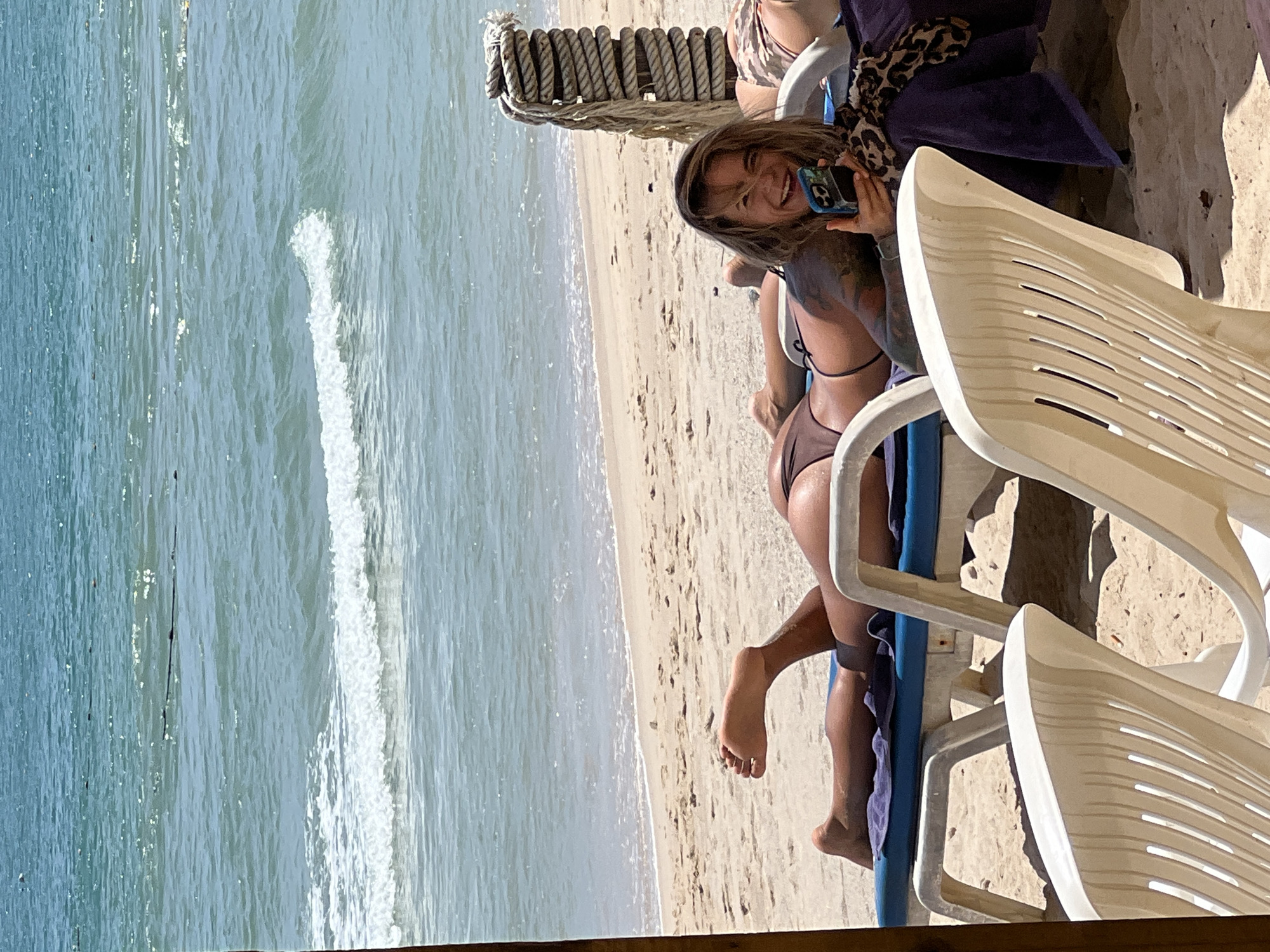 A woman lies on a beach chair on a sandy beach, facing the camera with a wide smile and holding a cell phone. She is wearing a brown bikini and has tattoos on her arms and shoulders. The ocean with gentle waves is visible behind her, and the sky is a clear blue. Two other beach chairs, one occupied by another person, are also in the frame. A thick rope is coiled around a post to the right of the woman. The scene appears to be outdoors in a tropical or warm climate.