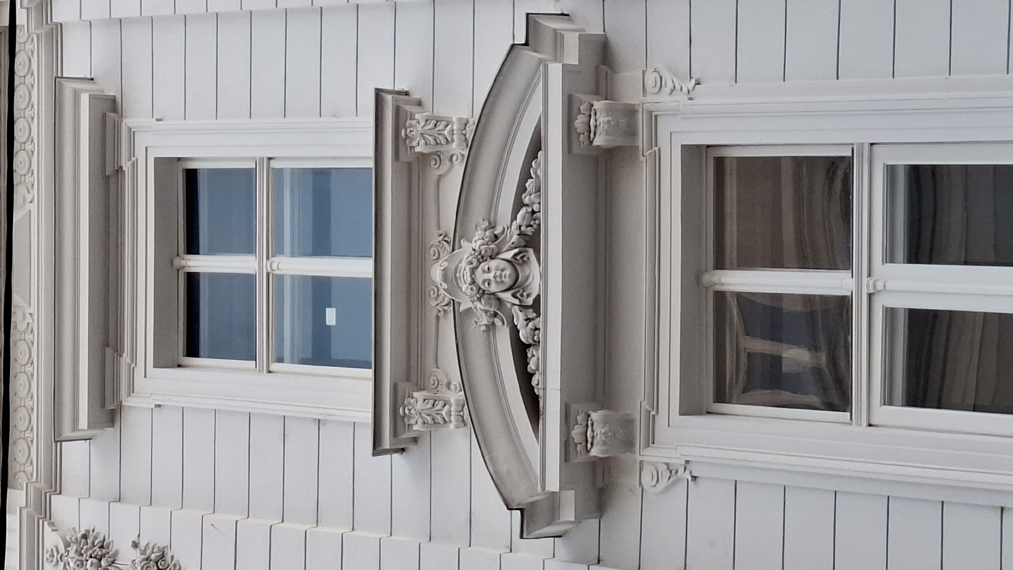 The image shows a close-up of a building facade in Vienna, Austria, featuring architectural details around a window. The building is painted white and has horizontal siding. There are two windows visible, one above the other. The upper window is framed by white molding and has a decorative arched element above it. This archway features a sculpted face with a floral headdress, flanked by ornate scrolls and architectural supports. The lower window is also framed by white molding and appears to have multiple panes. The image is taken from a slightly upward angle, focusing on the decorative elements and the windows.