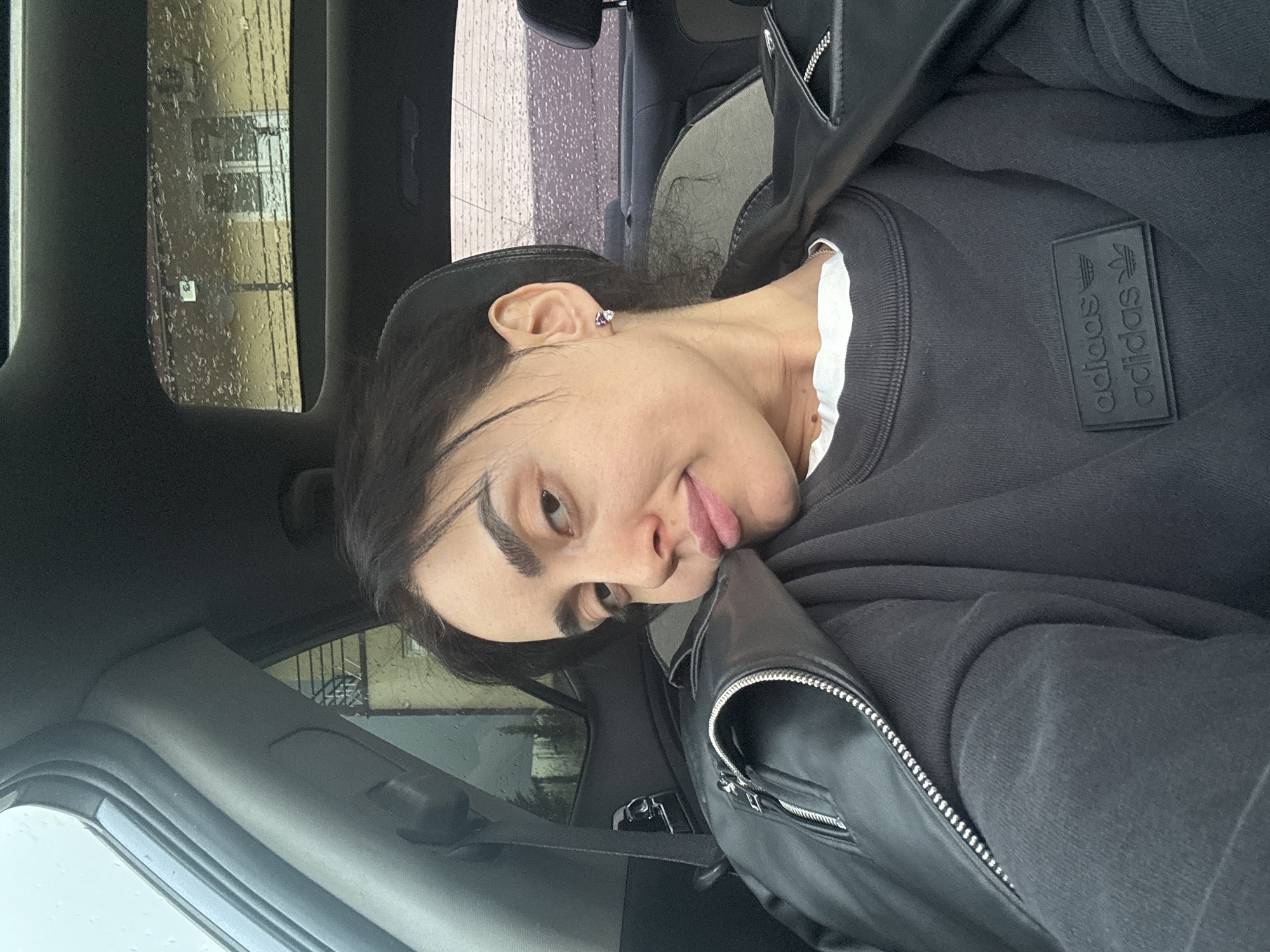 A person with dark hair and fair skin is in the driver's seat of a car. They are wearing a black Adidas sweatshirt and a black leather jacket. The car's interior is grey and black. Outside the car window, it appears to be a rainy day. There are buildings visible through the wet window. While a specific street address or landmark is not visible, and the image does not give enough information for definitive location, based on the architectural style that can be seen through the window, it is likely that the location of this image is Tbilisi, Georgia.
