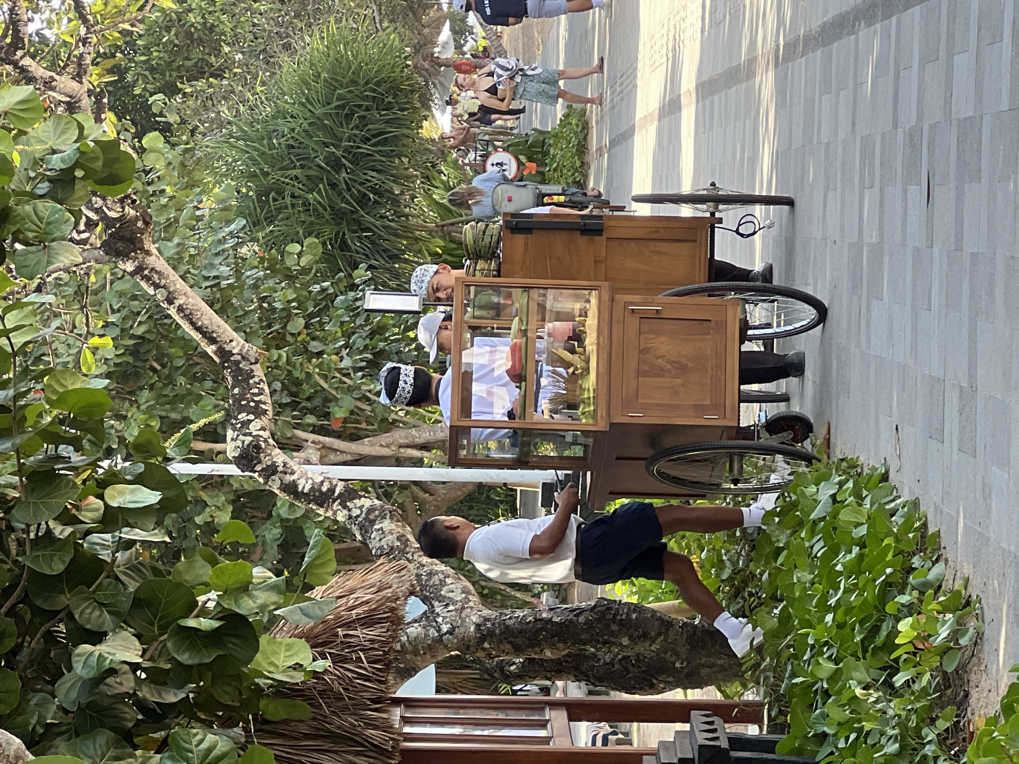 The image depicts a street scene in Denpasar, Indonesia, likely near a beach given the presence of palm trees and lush foliage. A wooden food cart with glass display cases is the central focus. Three individuals are interacting around the cart. One man, wearing a white polo shirt and dark shorts, is walking away from the cart, looking back. Two other men, dressed in traditional headwear, are standing near the cart, seemingly engaged in conversation or attending to it. The cart contains various food items, including what appear to be tropical fruits and possibly drinks in bottles. The ground is paved with light-colored tiles. In the background, other people are visible walking along the path, some with beach towels, suggesting a tourist area. The environment is outdoors, with dappled sunlight indicating a warm, sunny day.