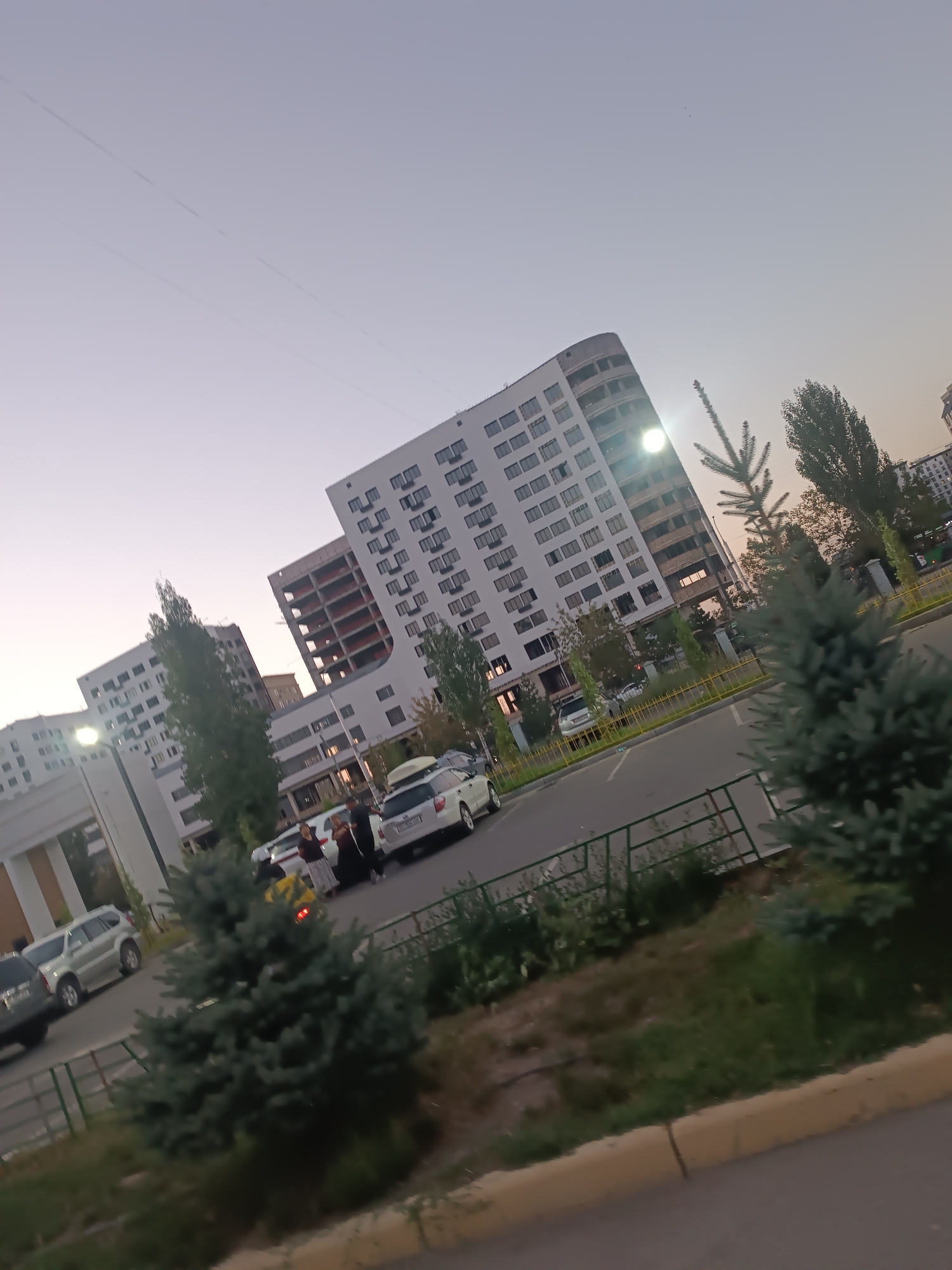 The image depicts a street scene in Bishkek, Kyrgyzstan, with modern apartment buildings dominating the background. The sky is a pale, hazy purple, suggesting either early morning or late evening. Several multi-story buildings, some with distinct curved facades and visible balconies, are present. One building appears to be under construction, with exposed red structural elements. In the foreground, a road is visible with parked cars, including a white SUV with a roof rack and a silver SUV. Several individuals are standing near the white SUV, seemingly interacting. Small, evergreen trees are scattered throughout the scene, adding some greenery. A metal fence with a yellow top runs along the edge of the road, separating it from a grassy area. The overall impression is of a residential area in a city.
