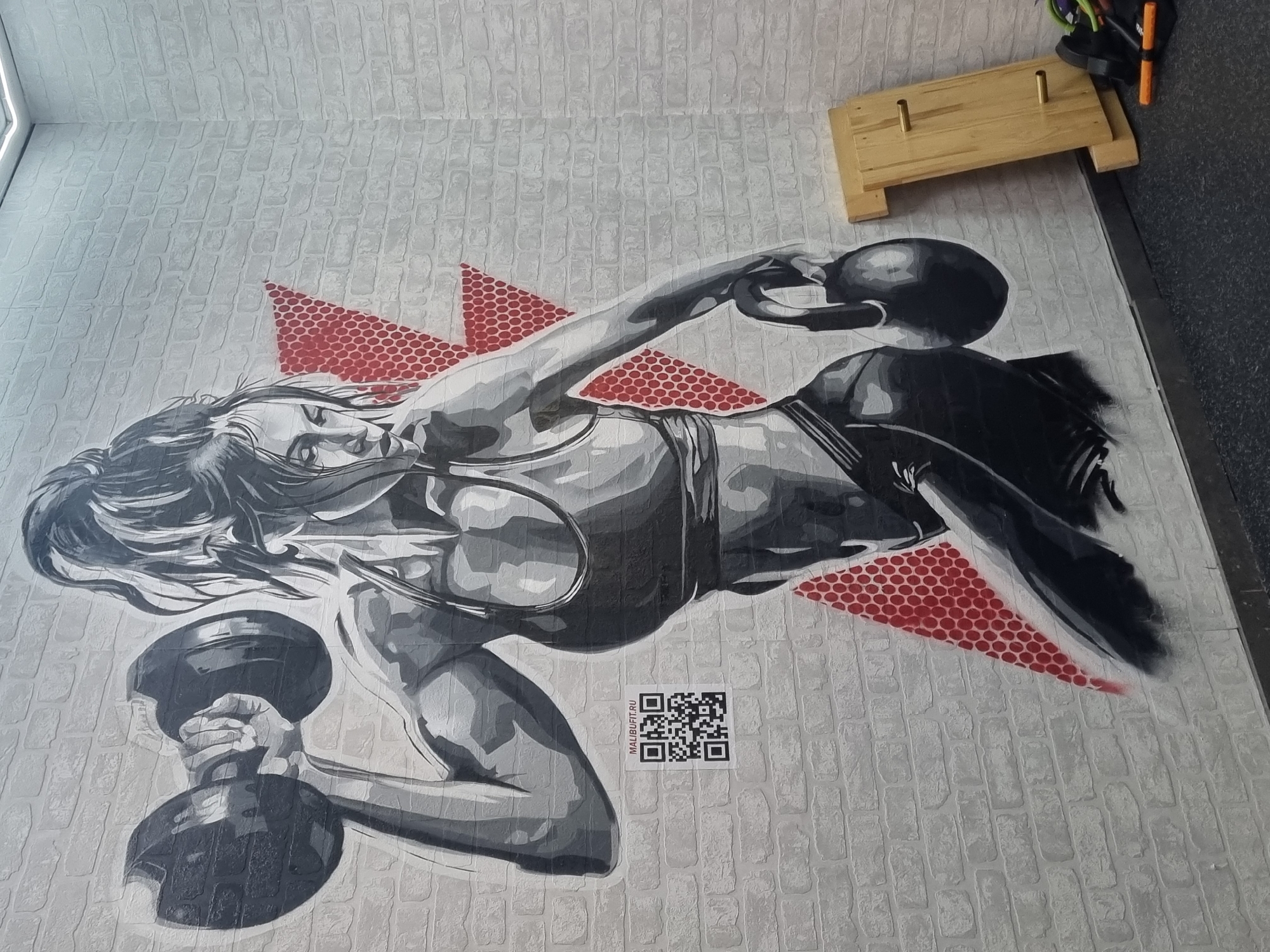 A floor-level perspective reveals a grayscale artwork of a muscular woman lifting kettlebells, set against a white brick-patterned floor. The artwork is punctuated by red, triangular shapes with a circular grid pattern. Adjacent to the artwork is a QR code and the text "MALIBUFIT.RU". In the upper right corner, a small wooden platform with metal posts is present, along with what appears to be a duffel bag. The scene is likely located in Sochi, Russia, inferred from the artwork's association with "MALIBUFIT.RU." The room where the artwork is displayed is possibly a gym or fitness studio.