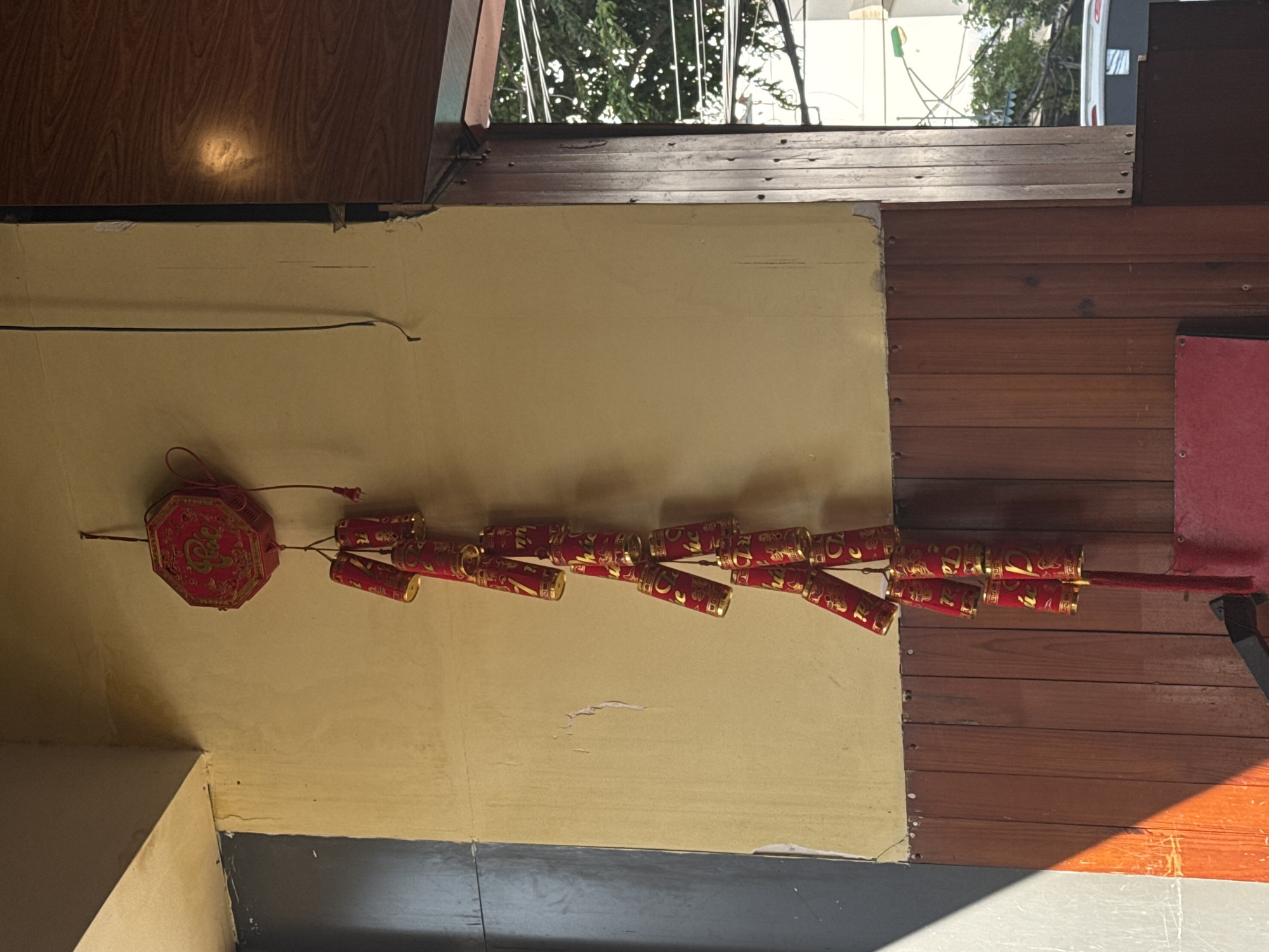 The image shows a string of Chinese firecrackers hanging from a pale yellow wall indoors. The firecrackers are red with gold accents and are connected to a decorative red ornament. The wall is patched and uneven, suggesting a slightly aged or weathered interior. To the right, the wall transitions to wooden planks. A small part of an open window is visible at the top, showing trees and buildings outside, suggesting this is an interior shot. The location is inferred to be in Nha Trang Ward, Vietnam, based on the prompt's request. There are no people visible, only the hanging firecrackers and the interior environment.