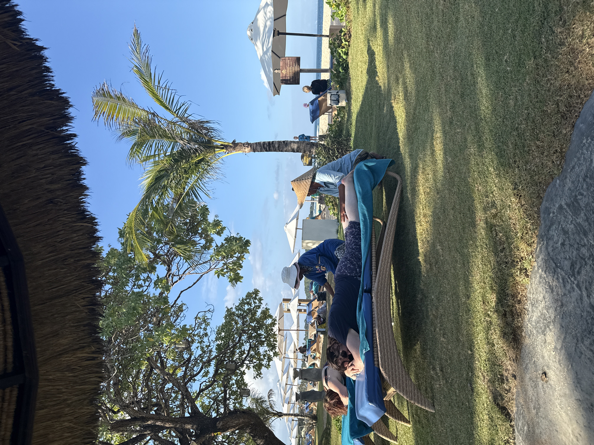The image depicts a scene at a beach resort in Kuta Selatan, Indonesia, under a thatched roof overhang. Two individuals are lying on lounge chairs on a grassy area, receiving massages from two therapists. Both therapists are wearing traditional conical hats. One therapist, wearing a blue shirt, is attending to the person in the foreground, who is lying on their stomach with their face turned to the side. The second therapist, in a light blue shirt, is working on the legs of another person on a neighboring lounge chair. In the background, several white umbrellas are set up on a sandy beach adjacent to the ocean. Palm trees and other lush vegetation are visible, and the sky is clear blue with some scattered clouds. The overall atmosphere suggests a relaxing resort experience.