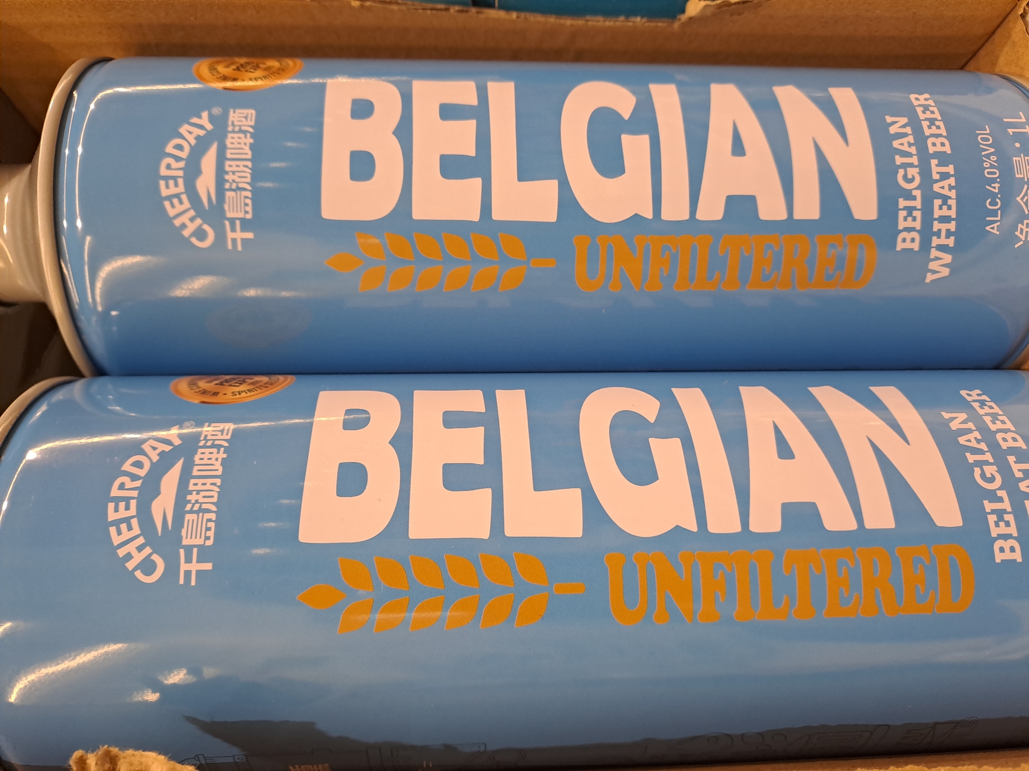 The image shows two cans of "CHEERDAY BELGIAN" wheat beer. The cans are light blue with white lettering for "BELGIAN", and gold lettering for "UNFILTERED" and "BELGIAN WHEAT BEER." The alcohol content is listed as 4.0% VOL and the volume is 1L. The brand name "CHEERDAY" and Chinese characters are also on the can. The cans are likely in a cardboard box. There is no mention of Elektrostal, Russia.