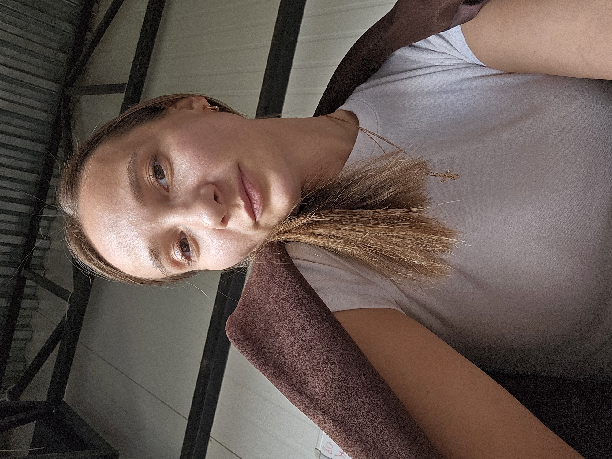 A light-skinned woman with brown hair is centered, looking directly at the camera with a slight tilt of her head. She is wearing a white t-shirt and a brown suede-like garment is partially visible over her shoulder. A thin gold necklace with a small pendant is visible around her neck. The background consists of corrugated metal and black metal framing, indicating an indoor or enclosed area. The location is Chaikovsky, Russia.