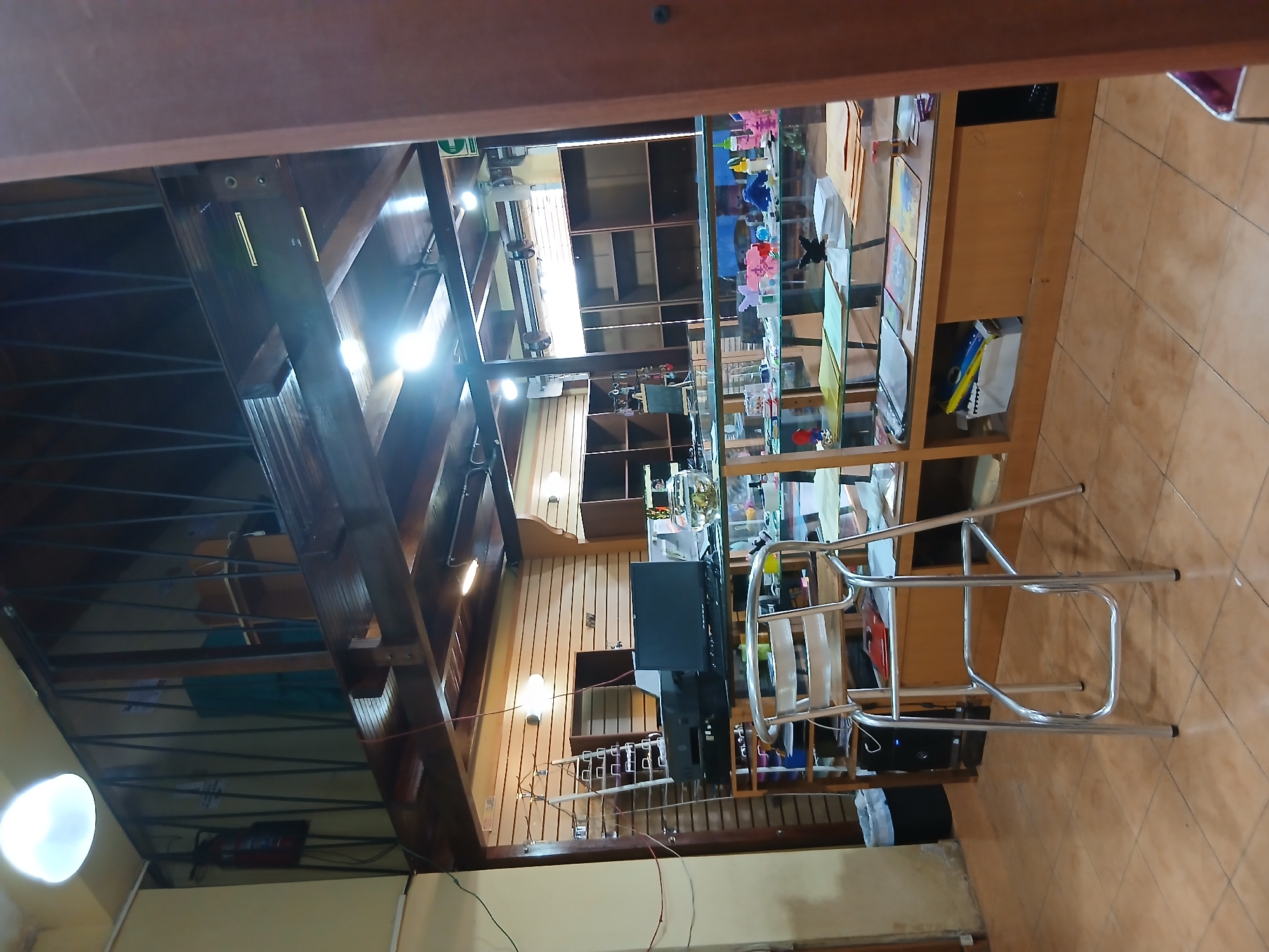 The image depicts the interior of a store in Juan Griego, Venezuela. The store features a mezzanine level with a dark wooden railing and a light fixture. Below the mezzanine, the main floor of the store is visible, with wooden paneled walls and a wooden ceiling with recessed lighting.
In the foreground, a silver metal chair with a rectangular seat and backrest is positioned on a tiled floor. To the left of the chair, a computer monitor, printer, and a black computer tower are situated on a counter. A fishbowl with some contents is also on the counter. A glass display case runs along the front of the counter, filled with various items including folders, boxes, and colorful objects. Behind the counter, there are shelves and cabinets.
To the left of the counter, a tall, silver ladder-like structure is leaning against the wall. A fire extinguisher is mounted on the wall near the mezzanine. The overall environment suggests a retail space, possibly selling stationery, craft supplies, or similar items. The lighting is a mix of overhead lights and natural light filtering from unseen sources. There are no people visible in the image.