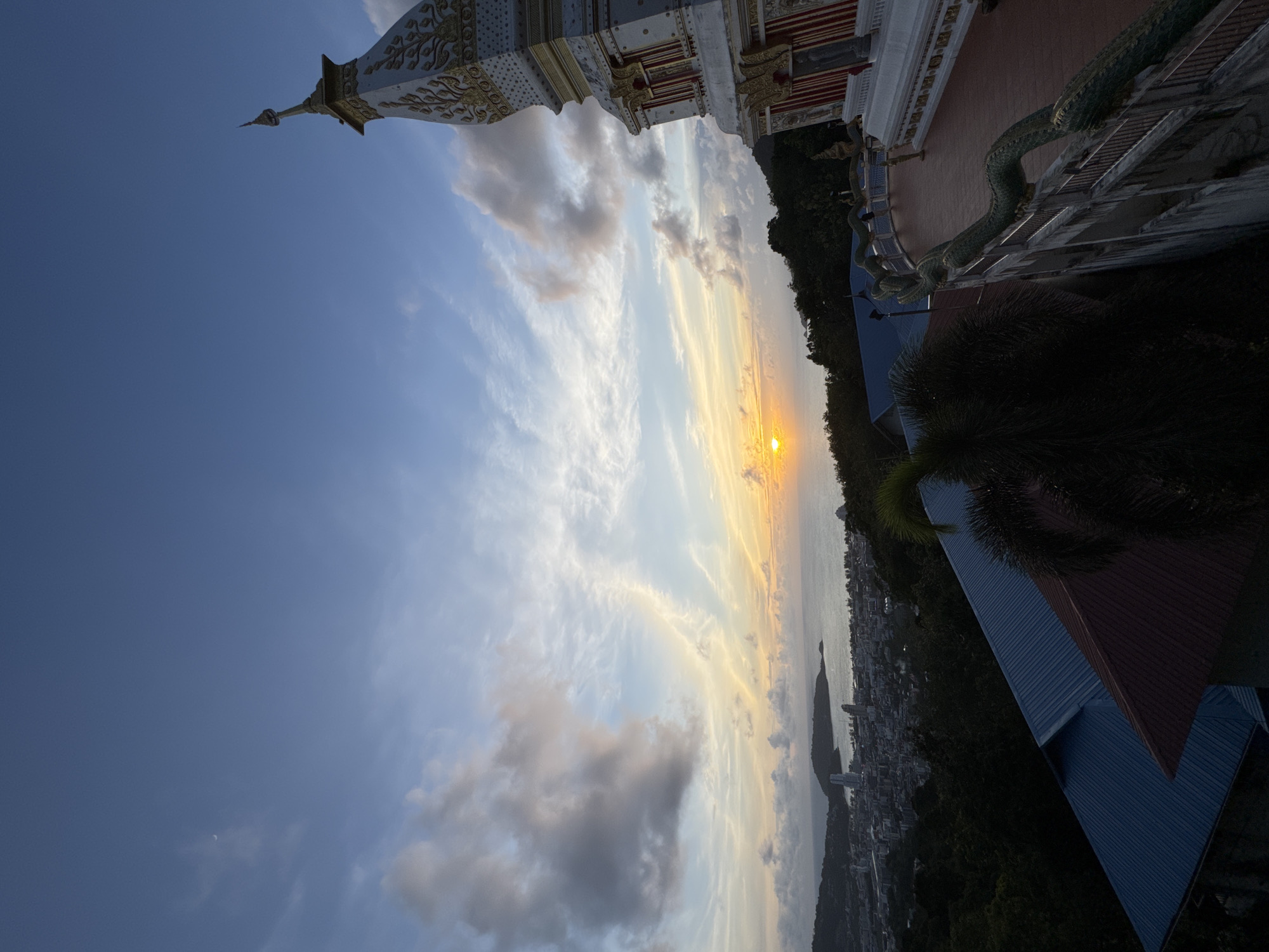 An elevated view of Patong, Thailand, captured at sunset, showcases the coastal cityscape and a Buddhist temple structure. The sun is low on the horizon, casting a warm glow over the sea and a crescent moon is visible in the sky. The temple, with its ornate white and gold stupa, dominates the right side of the frame, featuring intricate decorative elements and a naga-shaped railing along its balcony. In the foreground, the roofs of buildings are visible, one blue and one reddish-brown, nestled among dark green foliage. The background reveals the densely built-up city of Patong extending towards the coast, with the calm sea meeting the land. The sky is a blend of blue, white, and orange hues, with scattered clouds. The scene is tranquil and serene, typical of a tropical sunset.