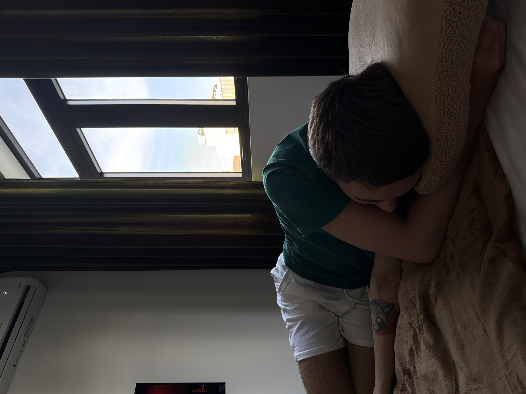 The image depicts a person, likely male, lying in bed with their face buried in a pillow. They are wearing a green t-shirt and white shorts. A tattoo of a circular design with a stylized animal or face is visible on their forearm. The setting appears to be a bedroom, with a window showing a bright sky and buildings outside. The window has horizontal blinds or slats and dark curtains frame it. An air conditioning unit is mounted on the wall to the left, displaying a digital temperature reading. The overall mood is one of rest or sleep, with the person seemingly seeking comfort in the pillow. The lighting suggests it is daytime, possibly morning or afternoon, given the bright natural light streaming through the window. The scene is domestic and relaxed. There is a glimpse of a television screen in the bottom left corner displaying a red graphic with text, but the text is not legible.