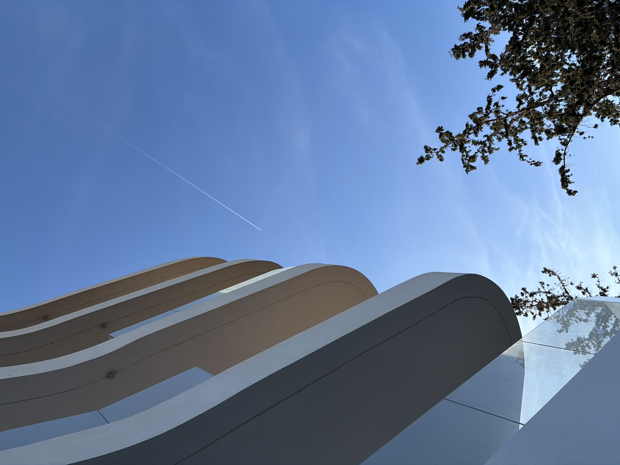 The image presents a low-angle view of a contemporary architectural structure, likely located in the Municipal Unit of Elliniko, Greece. The building features a series of curvilinear, cantilevered balconies or awnings, constructed from a smooth, likely concrete or composite material. The surfaces are light-colored, possibly white or a light grey, and reflect the ambient light. Clear glass panels are visible between the curved elements. The sky is a clear blue with light cirrus clouds and a visible contrail from a jet plane. A tree with dark foliage is positioned in the upper right corner of the frame, partially obscuring the sky. The photograph appears to be taken during daylight hours under sunny conditions.