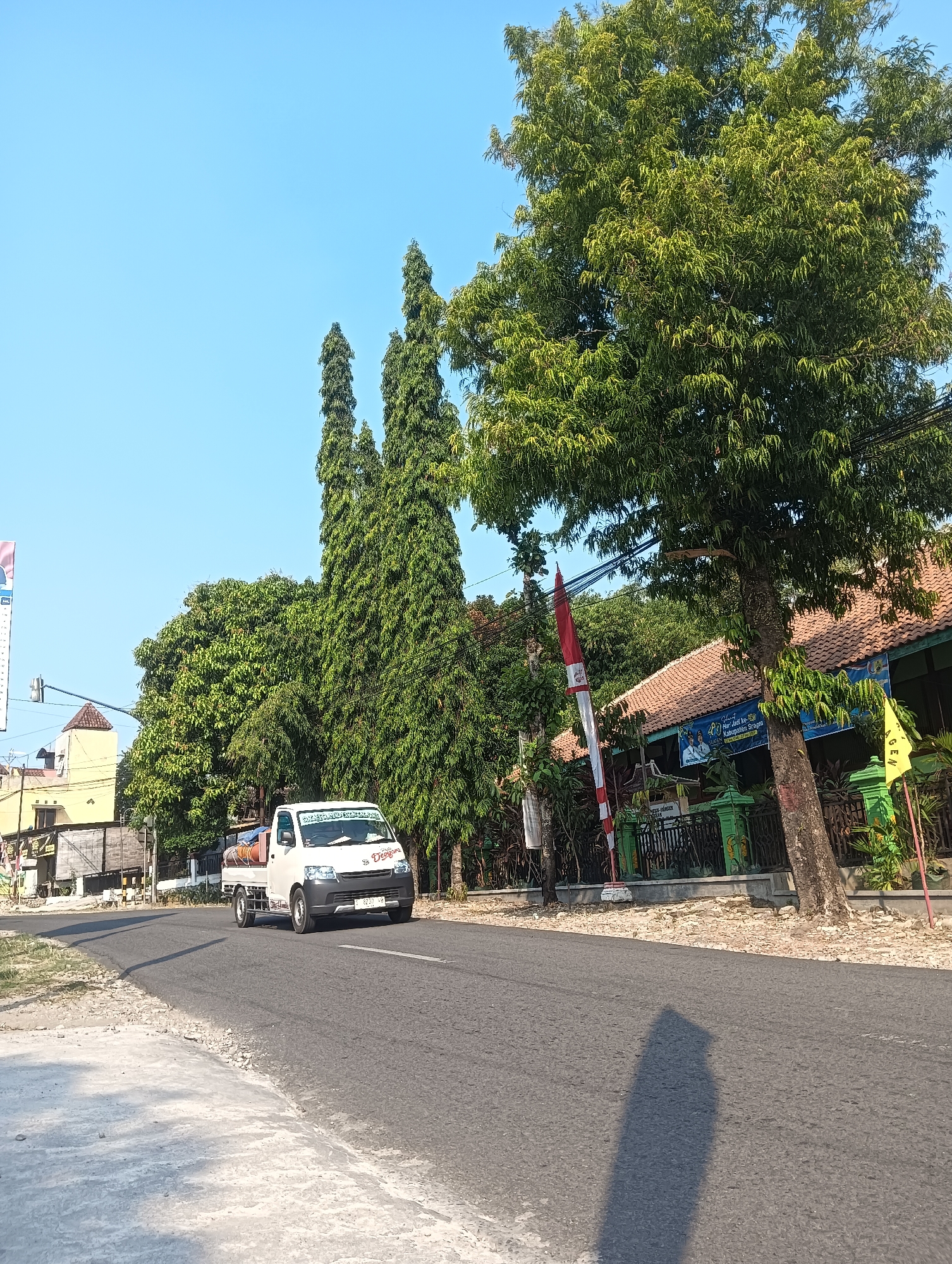 In Palemrejo, Indonesia, a white utility truck travels on an asphalt road on a clear, sunny day. The truck is carrying a cylindrical tank. On the left side of the road, a yellow building with a tiled roof is visible. A traffic light is mounted on a pole in front of the building. Tall, slender evergreen trees line the right side of the road, their foliage dense and green. A larger, mature tree with a thick trunk stands prominently on the right, its branches extending over the road. Behind the trees, a building with a terracotta tile roof and green trim is partially visible, adorned with banners and flags. A red and white striped flag and a yellow flag are planted near the road. The asphalt road curves gently to the left, with a concrete sidewalk on the left and a gravel shoulder on the right. The sun casts a long shadow of a tall object, likely a tree or pole, across the road.