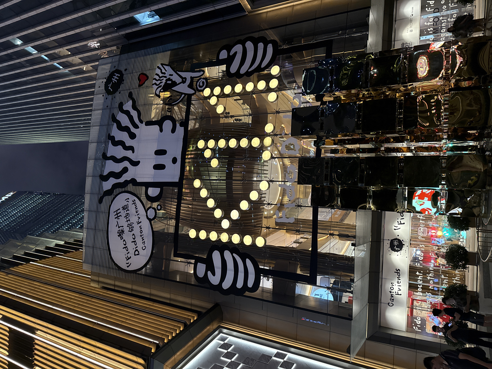 The image depicts a nighttime exterior view of a modern commercial building, likely a shopping mall or department store, located in Tianhe District, China. The building features extensive glass facades illuminated by various lighting elements. Prominently displayed on the upper floors is a large graphic of a stylized cartoon character with black and white stripes, holding a red heart and a black hand with white fingers. A speech bubble above the character contains Chinese characters and the English text "Fido Guangzhou Dido 够地道 Canton Friends". The building's lower section has a storefront with the text "Canton Friends" and further text in English that appears to be advertising slogans. In front of the building, several individuals are visible, some standing and looking at their phones, while others are walking. A reflective, abstract sculpture made of dark, multi-faceted blocks stands in the foreground, casting distorted reflections of the surrounding lights and buildings. The overall scene conveys a vibrant, urban nighttime atmosphere.