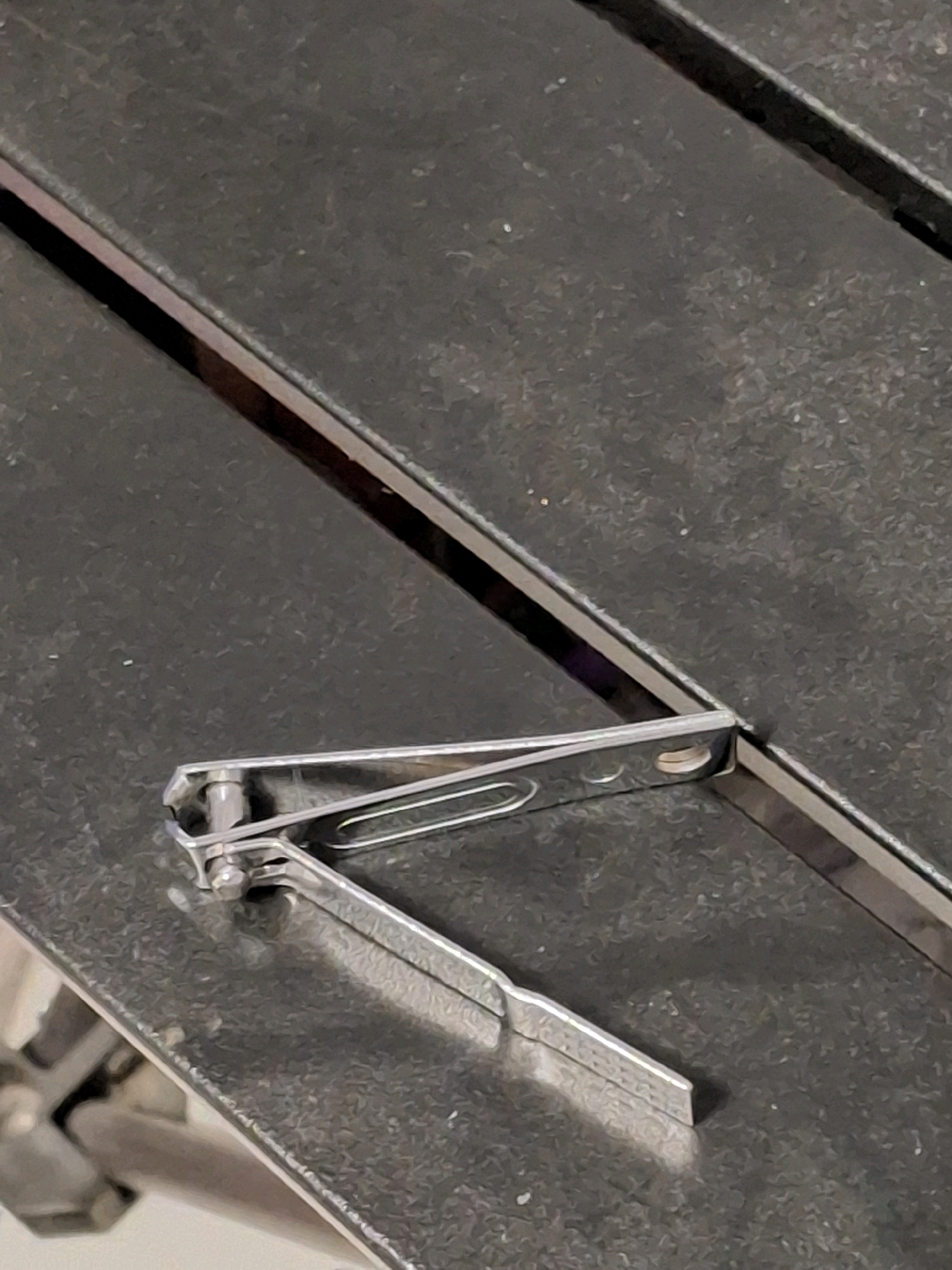 A pair of silver nail clippers rests on a dark, textured surface. The nail clippers are partially open, with the lever bent upwards and resting on the other half of the tool. The surface appears to be a table or countertop, with a faint, dark line running diagonally across it behind the clippers. The lighting is somewhat dim, casting a slight shadow from the nail clippers. No people are present in the image. The location in Lumajang, Indonesia, is not discernible from the visual information provided.