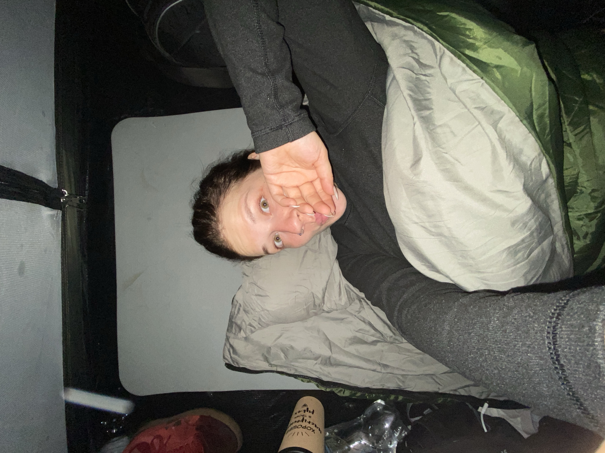 A young woman is lying in a sleeping bag inside a tent. She is wearing a dark gray long-sleeved shirt and dark gray pants. Her hair is dark brown and pulled back. She is looking at the camera with her mouth slightly open and her right hand covering her mouth. There is a white pillow under her head. To the left of the sleeping bag is a gray mat, and to the left of that is a red shoe and a brown cylindrical object with writing on it. The tent appears to be set up in an outdoor location. The image was taken in Абзаново, Russia.