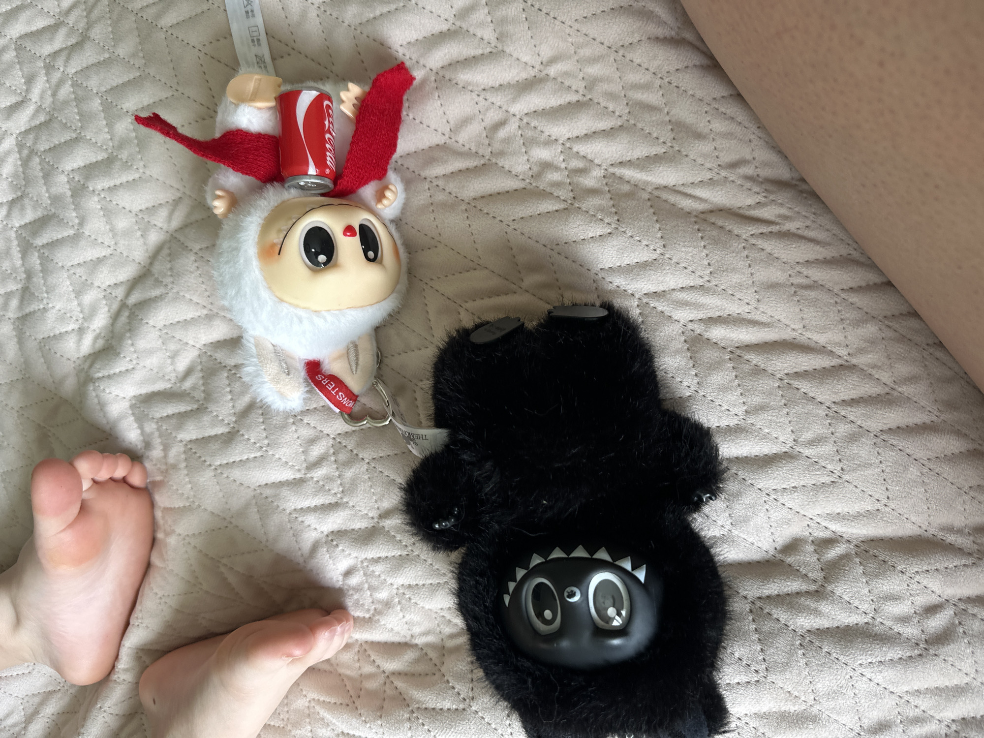 The image shows a bed with two plush toys. On the left is a white toy with a red Coca-Cola can on its head, red scarf and red accents around the head. On the right is a black furry monster toy with a black plastic face with white teeth. There are also two human feet at the bottom left and a leg in the top right corner of the image. The bed has a light beige quilted pattern. The toys are placed on the bed. The location of Chaikovsky, Russia is unknown.