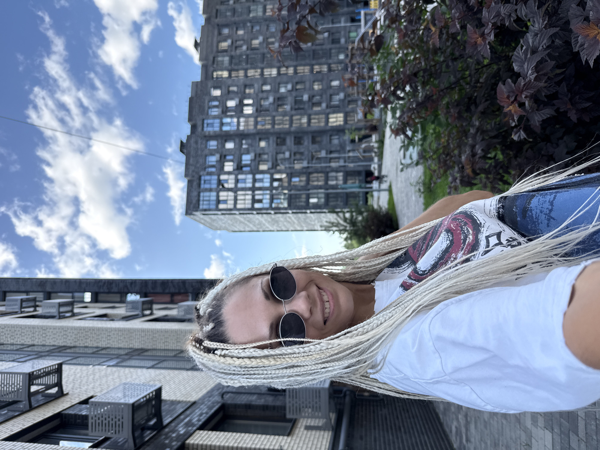 A young woman with blonde braids and sunglasses is taking a selfie in front of apartment buildings and greenery. The woman is wearing a white t-shirt. The buildings are modern and multi-story, with windows visible. The sky is blue with scattered clouds. The ground is paved with bricks, and there are some bushes and trees in the foreground and background. The overall scene suggests an urban outdoor setting. The location is identified as Rusanovo, Russia.