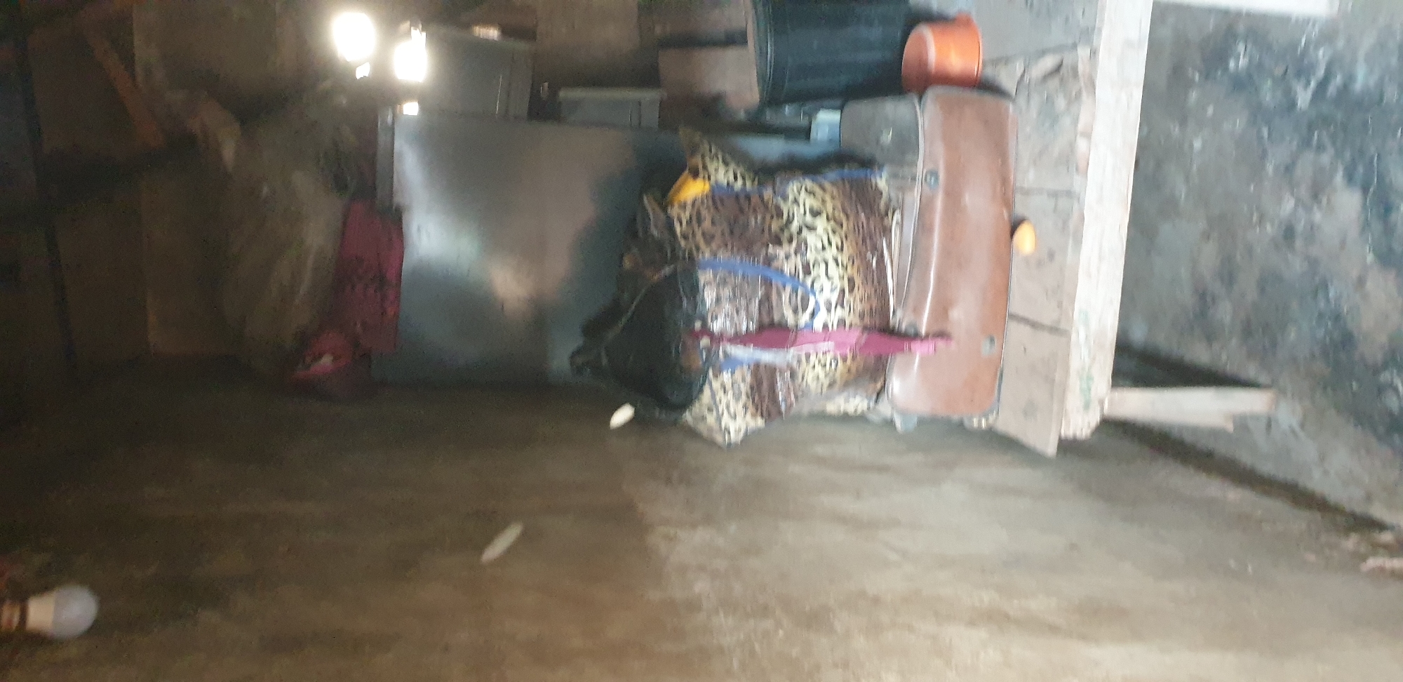 The image depicts an indoor scene, possibly a storage area or a less-maintained room in Sabon Tasha, Nigeria. The walls are rough and unpainted, suggesting a rustic or utilitarian environment. On the left, a lightbulb hangs from the ceiling, illuminating the space with a diffused light.
In the center of the image, a collection of items is piled on a wooden table or shelf. Dominating this collection is a large, leopard-print bag, secured with blue and red straps. A black object, possibly a hat or another bag, rests on top of the leopard-print bag. Beneath this, a brown, flat object, resembling a briefcase or a piece of luggage, is also visible. An orange bucket and a black container are situated to the right of the table, suggesting storage or waste disposal.
The floor appears to be made of concrete or compacted earth, with visible debris. The overall impression is one of a functional, perhaps cluttered, space, likely within a residential or workshop setting in the Sabon Tasha area. No individuals are present in the frame.