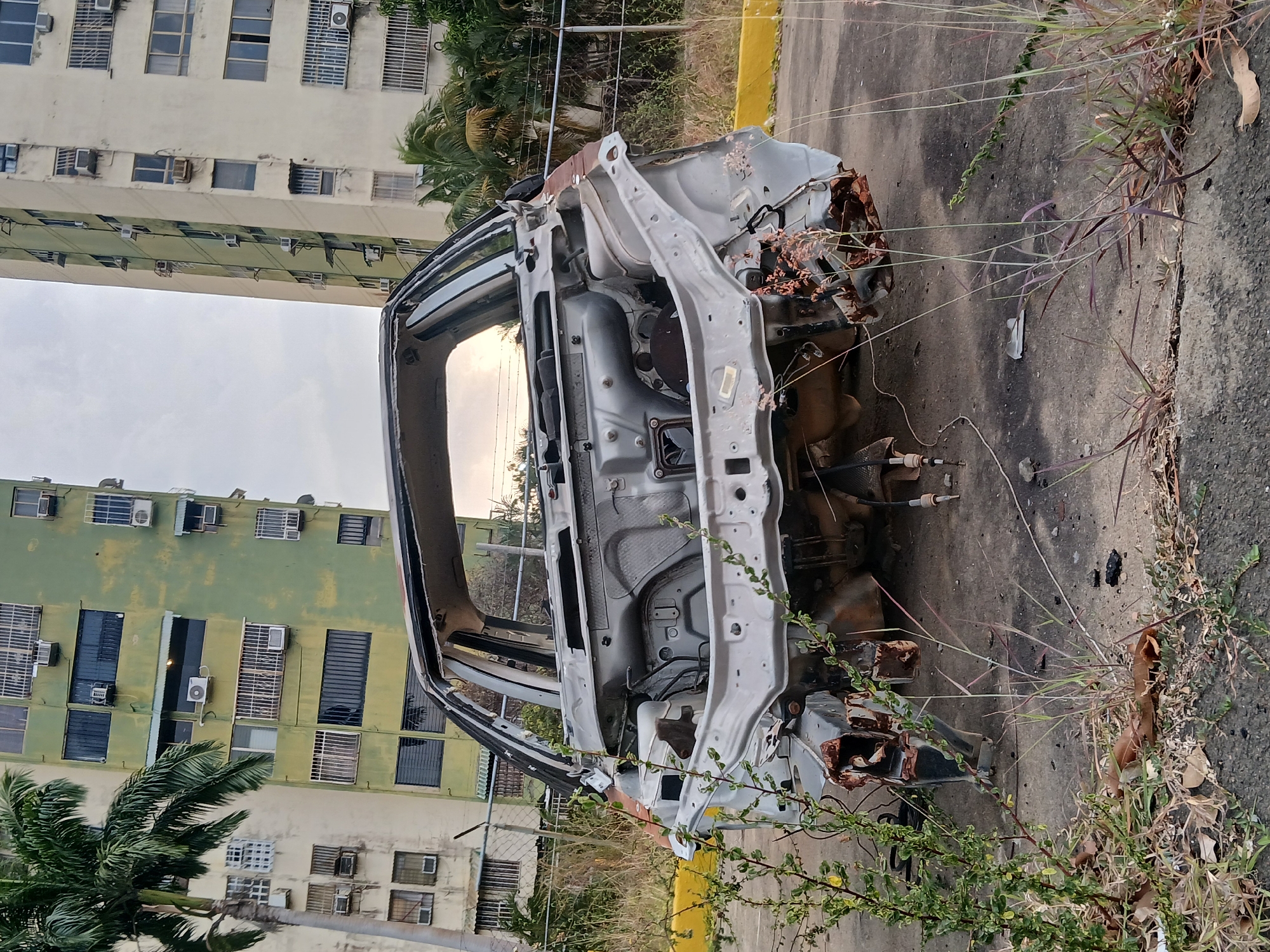 The image captures a scene of urban decay featuring the skeletal remains of a stripped car chassis, lying derelict on an asphalt or concrete surface. The car, likely a sedan or hatchback, has been almost entirely scavenged, with its body panels, interior, engine, and wheels missing, leaving only the rust-streaked, light-colored metal frame. Rust is particularly visible on the front and rear sections where components would have once been attached.
Around the abandoned vehicle, the ground shows signs of neglect, with weeds and tall grass growing through cracks in the pavement and along a yellow-painted curb. Various pieces of discarded debris are scattered near the car. Palm trees and other lush green foliage are visible in the midground and background, indicating a tropical or subtropical climate.
In the background, multiple tall, multi-story residential buildings rise against a hazy or overcast sky. These buildings appear somewhat aged, with visible signs of wear and numerous external air conditioning units attached to their facades. One building has a distinct light green hue, while another is a paler beige or cream color. A chain-link fence can be seen separating the foreground area from the buildings and other properties.
The scene depicts the aftermath of abandonment and scavenging, with the stationary car serving as a stark reminder of neglect. The time of day appears to be daytime, with soft, diffused light suggesting either early morning or late afternoon, or an overcast sky. The overall atmosphere conveys a sense of desolation and urban blight, consistent with areas experiencing economic challenges, as might be found in Ciudad Guayana, Venezuela. No visible text is present in the image.