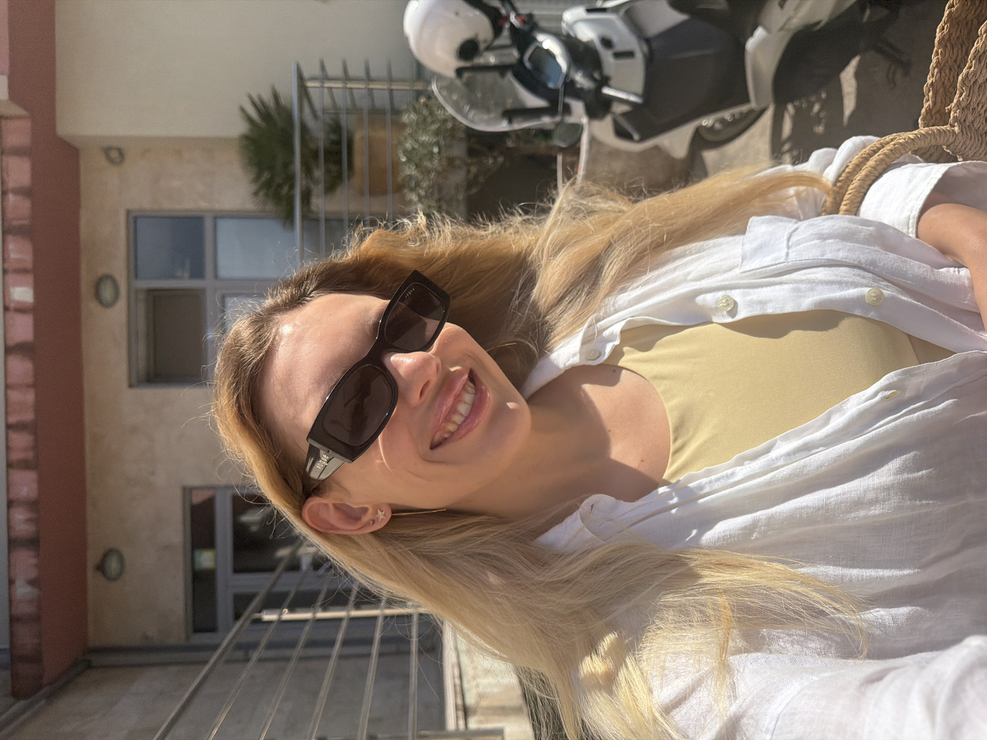 A young woman with blonde hair, wearing sunglasses and a white open shirt over a yellow top, smiles at the camera. She is outdoors, with a building and a scooter in the background. The building appears to be made of light-colored stone with a red-tiled roof and balconies with metal railings. A potted plant is visible on one of the balconies. The scooter is white with a black seat and a windshield. The scene suggests a sunny day. The location is Budva, Montenegro.