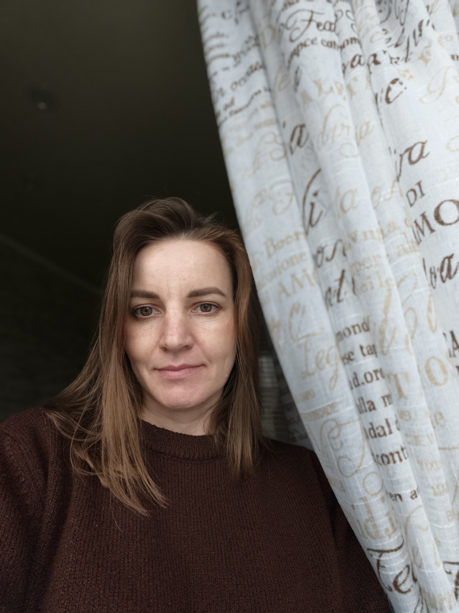 A portrait of a middle-aged woman with light brown hair, wearing a brown sweater, stands indoors next to a light-colored curtain printed with script and Italian text. The background is dark, suggesting a dim room or window. The woman is looking directly at the camera with a neutral expression. The location is not specified in the image.