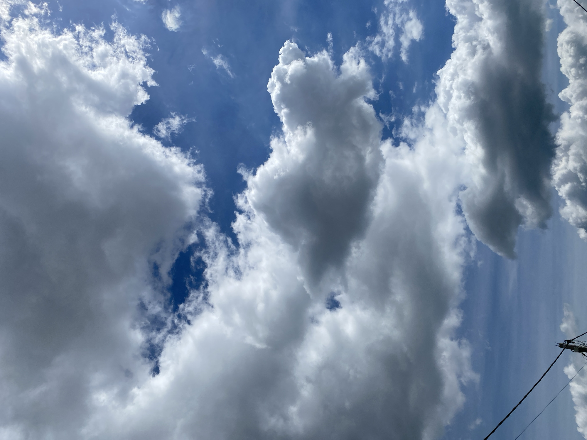 The image depicts a sky filled with a mix of white and gray clouds against a bright blue background. The clouds vary in size and density, with some appearing fluffy and illuminated by sunlight, while others are darker and more ominous. In the lower portion of the image, a black wire runs diagonally from the left, and a utility pole with wires is visible, indicating an urban or suburban setting. The scene is devoid of any people or specific landmarks that would pinpoint a city or location.