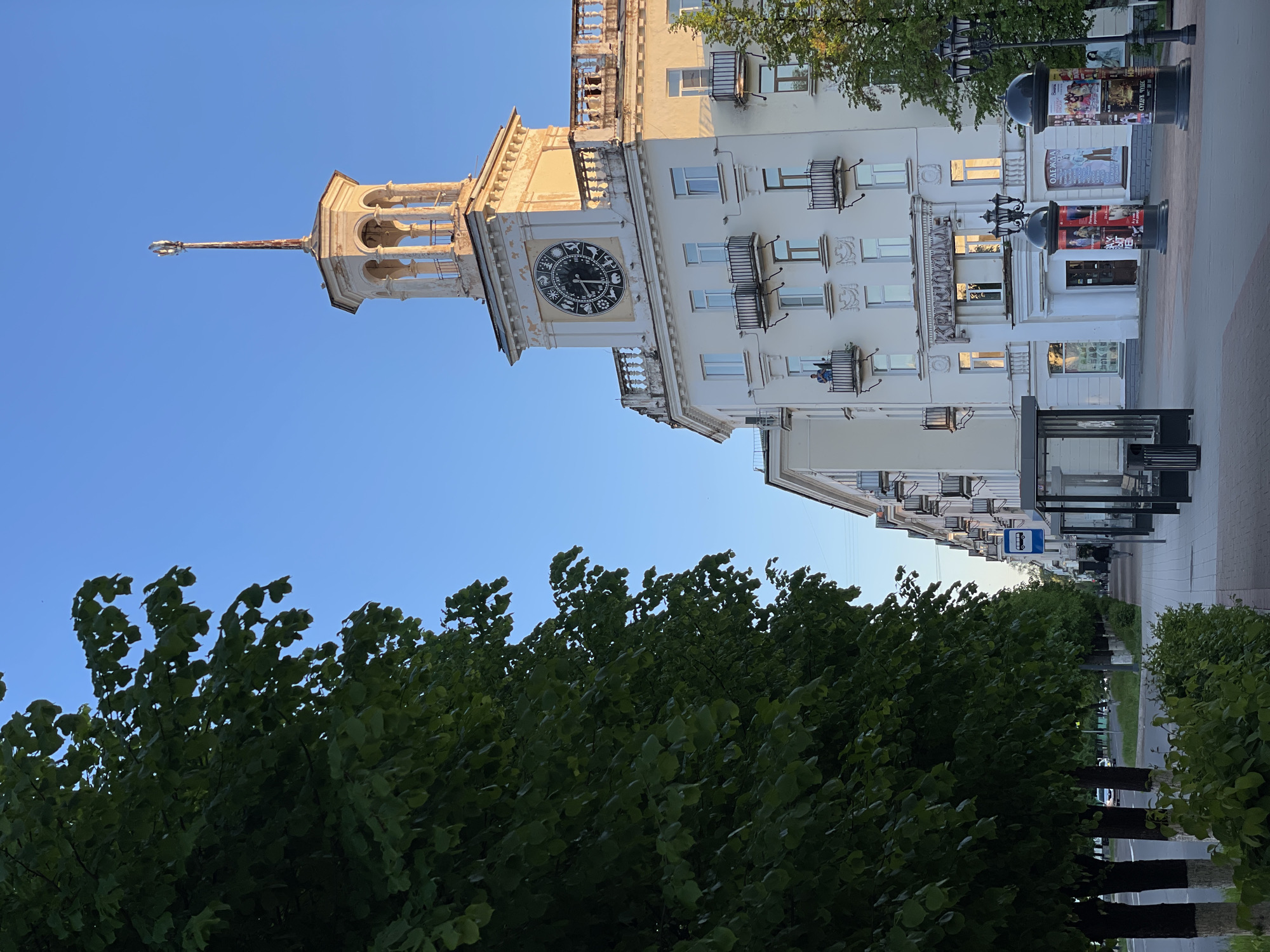 The image shows a street scene in Zheleznogorsk, Russia, featuring a prominent building with a clock tower. The building is white with multiple windows and small balconies. The clock tower has a spire extending upwards. In the foreground, there are trees with green foliage. A sidewalk runs along the base of the building. Several black street poles with posters are visible on the sidewalk. A bus stop is also present. The sky above is clear and blue.