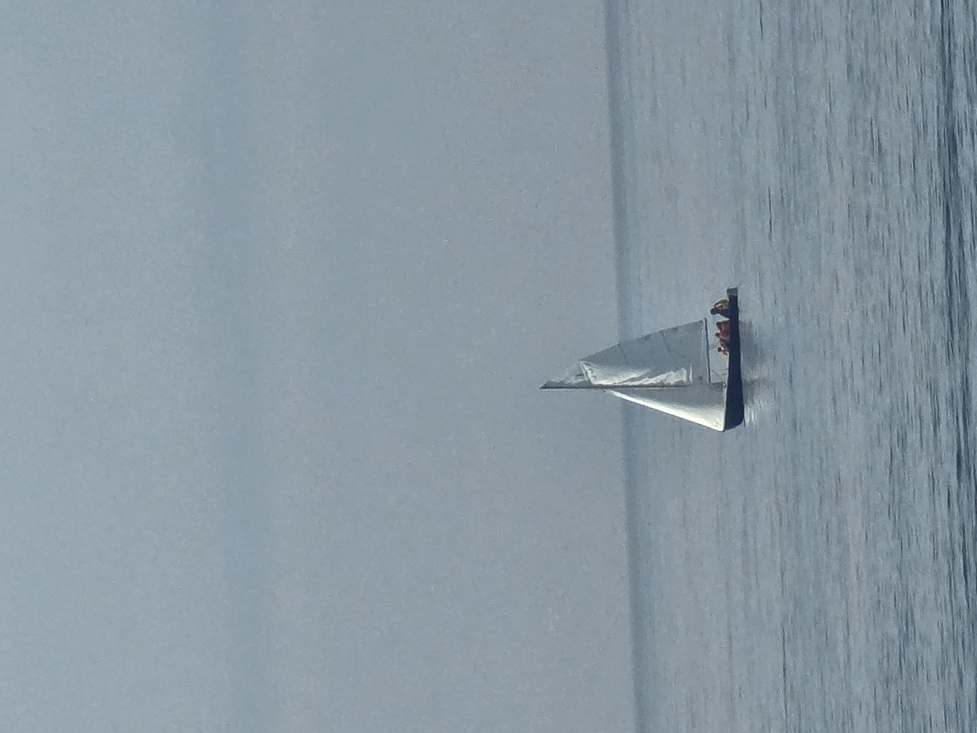The image depicts a solitary sailboat on calm, blue water under a hazy, overcast sky. The sailboat, with a white sail, is positioned in the center-right of the frame. Four individuals, wearing orange life vests, are visible on the deck of the sailboat. The sea is tranquil, with subtle ripples on its surface. The background is dominated by the muted tones of the sky, creating a serene and slightly diffused atmosphere. This scene could be occurring anywhere along the coast of Málaga, Spain, given the common presence of sailboats and the typical coastal weather conditions.