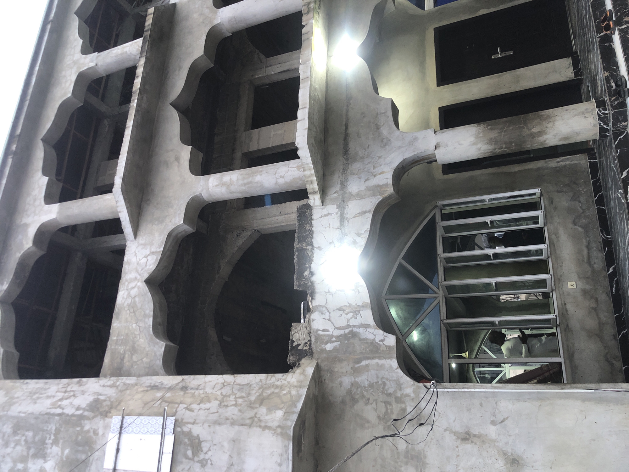 This image depicts the unfinished facade of a concrete building, characterized by its multiple levels of arched openings and columns. The building's exterior shows signs of age and incomplete construction, with visible cracks and staining on the concrete surfaces. On the ground level, a large window with multiple panes is visible, and through it, one can see at least two individuals dressed in white attire, suggesting a religious or cultural context. One individual appears to be holding something in their hands, possibly in prayer or contemplation. Two dark doors are situated to the right of the window, with a pair of sandals placed on the tiled ground in front of them. The architecture suggests a location in Ado, Nigeria, potentially a mosque or community center in the process of construction or renovation.