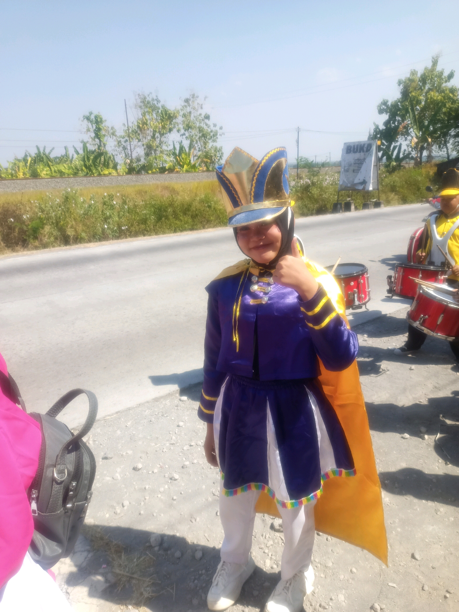 In Sumberlawang, Indonesia, a young person wearing a purple and gold marching band uniform, complete with a distinctive helmet, gives a thumbs-up to the camera. They are standing outdoors on a paved road, with a grassy area and trees visible in the background. Another individual in a yellow uniform, holding drumsticks, is partially visible in the background, along with red drums. A black backpack rests on the ground to the left of the frame. The scene suggests a warm, sunny day, likely during a parade or performance.