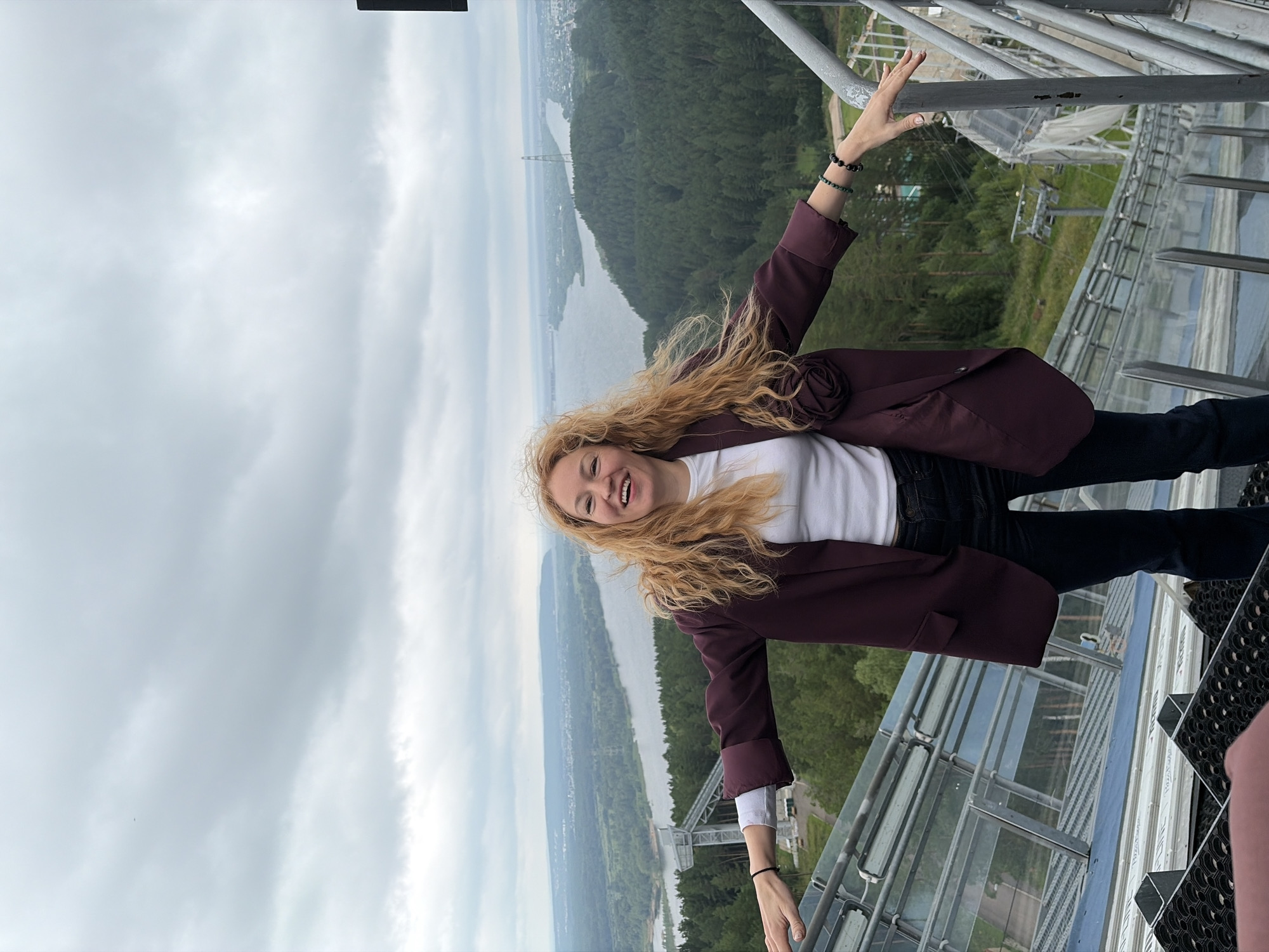 A woman with long, wavy blonde hair stands with her arms outstretched, smiling at the camera. She is wearing a dark burgundy blazer over a white shirt and dark jeans. She is standing on a metal structure with glass panels. In the background, there is a wide river, a forest, and a city skyline under a cloudy sky. The location is identified as the trampline of the "Lysovka" ski jump complex in Perm, Russia, often referred to as part of the "Prikamsky" region due to its proximity to the Kama River.