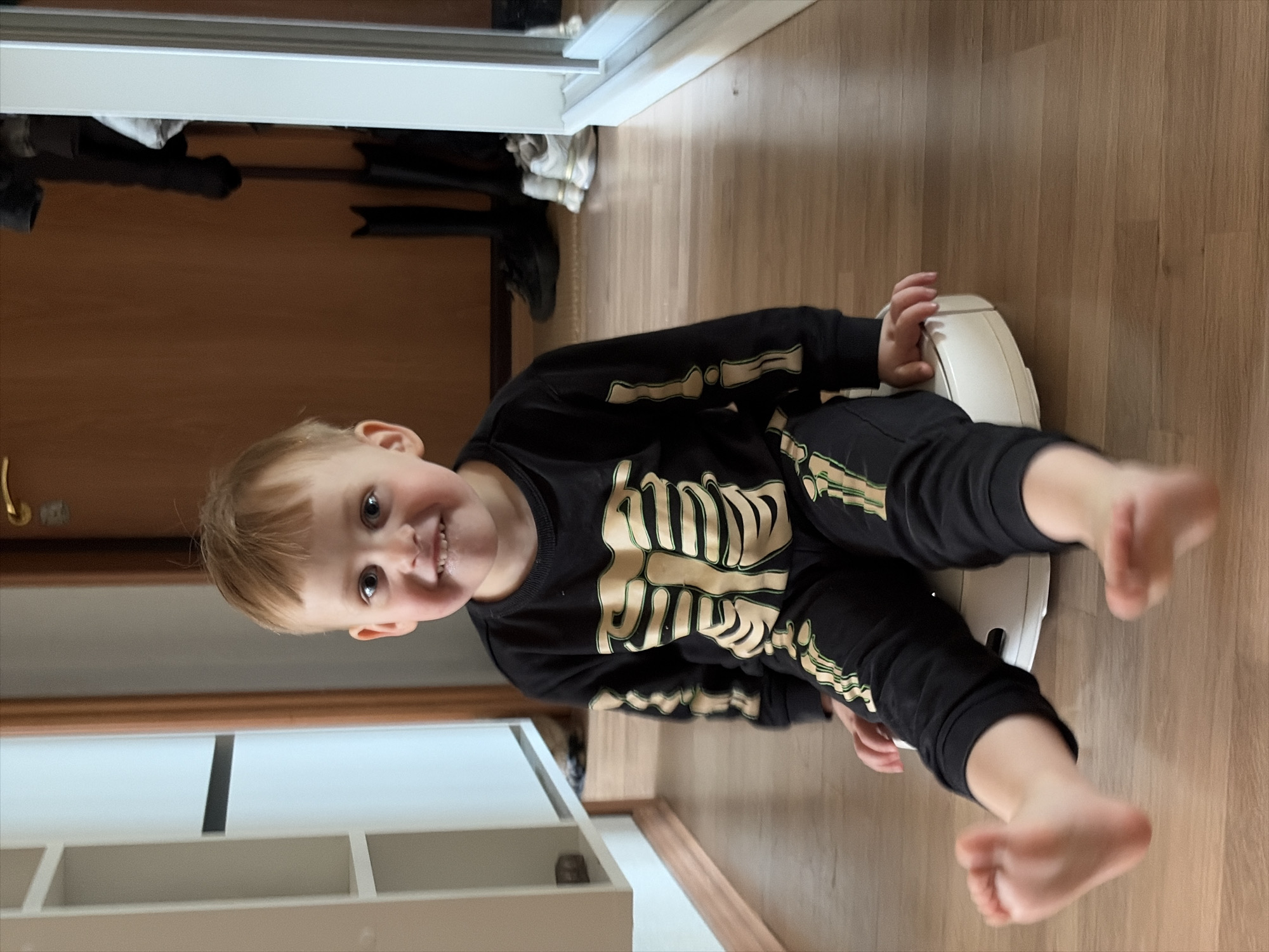 A young child in a black skeleton outfit sits cross-legged on a white robotic vacuum on a wood floor. The child is facing forward with a small smile and their feet are extended in the air. A white storage unit and a wooden door are visible in the background. The location is unknown, and there is no indication it is Chaikovsky, Russia.