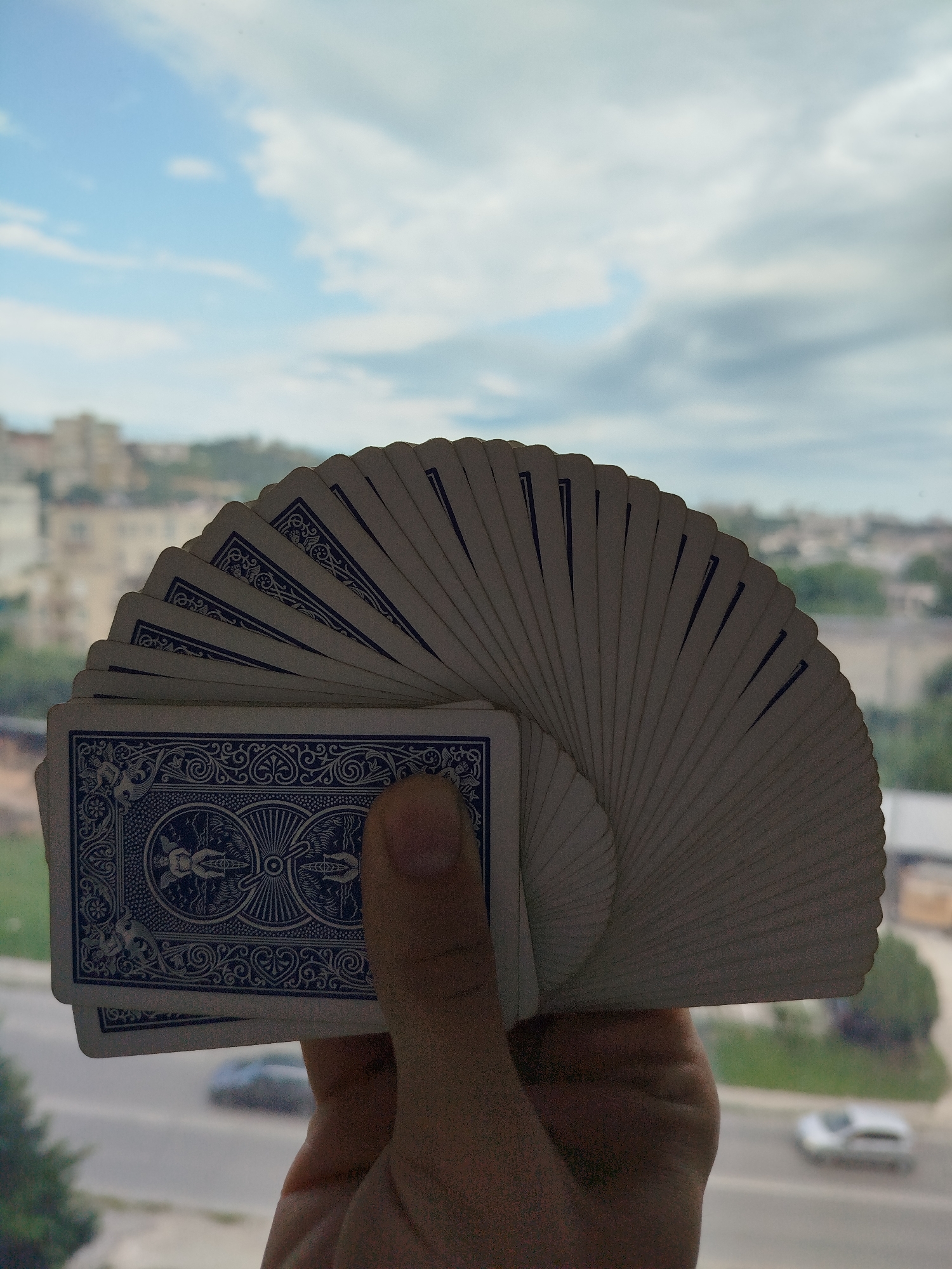 A hand holds a fan of blue-backed playing cards. The backs of the cards are visible, featuring the Bicycle brand design. The background shows an urban landscape with buildings and greenery under a cloudy sky. Based on the architecture and environment, the location is likely Varna, Bulgaria. Cars are visible on a road in the background.