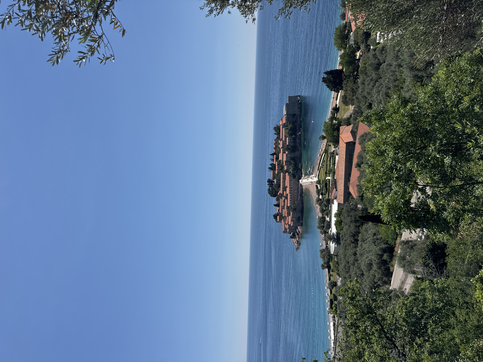 The image captures a coastal view in Sveti Stefan, Montenegro. The Adriatic Sea stretches towards the horizon under a clear blue sky. In the center of the image is Sveti Stefan island, a small island resort with distinctive terracotta rooftops. The coastline is visible to the right, marked by a strip of land with trees and some buildings also with terracotta roofs. The view is framed by tree branches in the upper corners of the image.