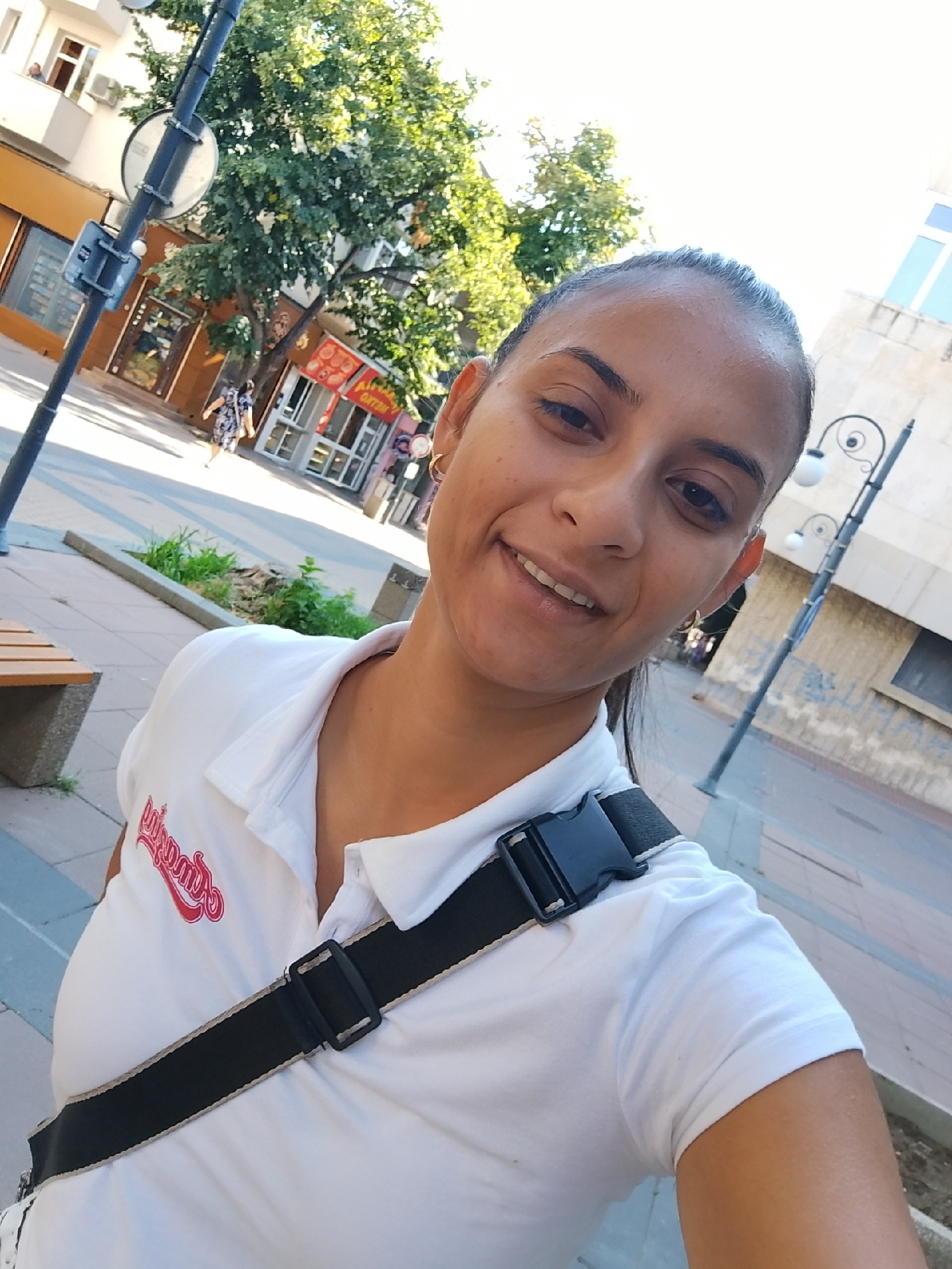 The image is a selfie taken outdoors in the city of Yambol, Bulgaria. The subject, a woman with dark hair and a wide smile, is wearing a white polo shirt with a red logo and a black strap across her chest. In the background, there are buildings, trees, a lamppost, and a bench. The scene appears to be a pedestrian area or a street in a town. The lighting suggests it is daytime.