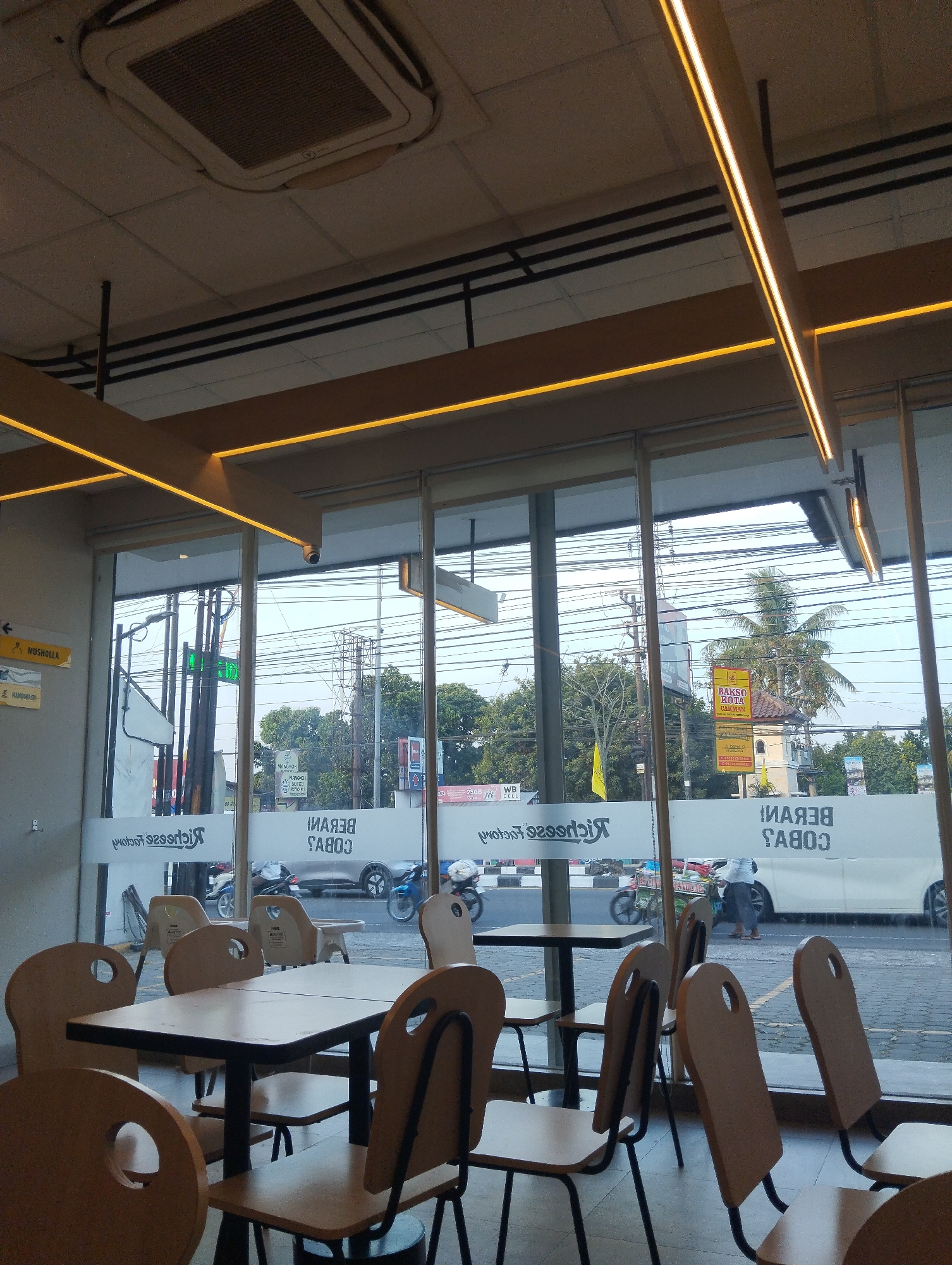 The image depicts the interior of a restaurant, likely a fast-food establishment, viewed through a large window. The restaurant's branding, "Richchfise Factory" is visible on the glass in reverse. Several tables and chairs are arranged inside. Outside, a street scene is visible with various vehicles, including motorcycles and cars, as well as utility poles and trees. A sign for "BAKSO KOTA CAKMAN" is prominent. The overall lighting suggests it is daytime. The presence of the street and typical Indonesian signage indicates this is likely a commercial area in Sumberrejo, Indonesia.