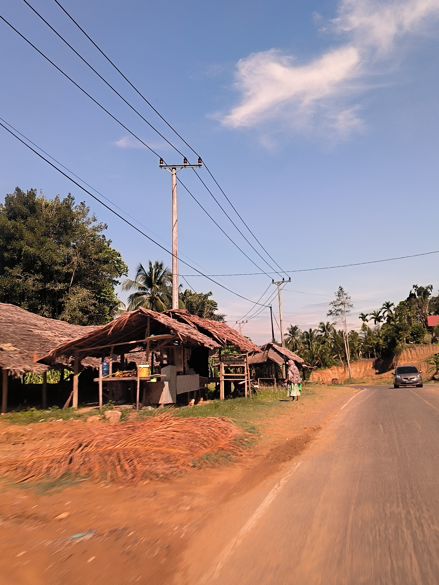 The image presents a sun-drenched roadside scene in Tanjongkeumala, Indonesia, under a clear blue sky dotted with a few wispy white clouds. On the left side of the frame, several rustic, open-air stalls or huts with traditional thatched roofs, constructed from dried palm fronds, line the dirt shoulder. One stall features a wooden table holding a large bottled water container, possibly some fruit, and a yellow bucket, suggesting it's a small shop or food stand.
In front of these structures, large piles of dried reddish-brown palm fronds are scattered on the ground. Further along the roadside, near another hut, a person, likely a woman, stands wearing a head covering and a patterned long dress or skirt over a green garment.
Behind the stalls and along the elevated terrain to the right, a dense canopy of lush tropical vegetation, including tall trees and numerous palm trees, characterizes the landscape. Several utility poles with power lines stretch across the sky, indicating a degree of infrastructure in the area. The ground on the right side of the road shows exposed reddish-brown soil on a gentle slope.
An asphalt road, appearing to be a two-lane thoroughfare, curves into the distance on the right. A silver or grey car is visible driving along this road, moving away from the viewer. The bright light and distinct shadows suggest it is either morning or early afternoon. The scene conveys a calm, everyday rural atmosphere. No visible text is present in the image.