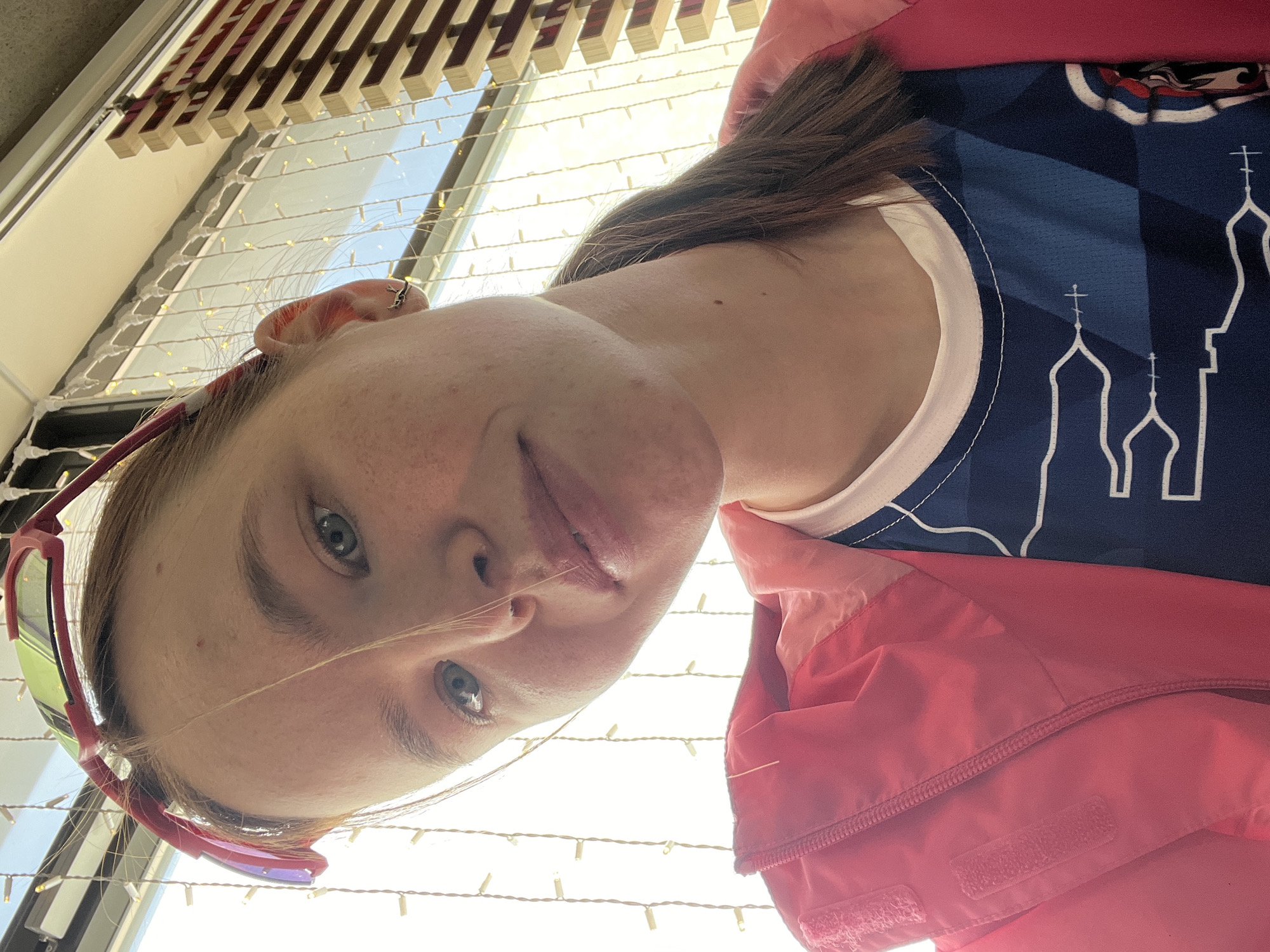 A young woman with fair skin and freckles is facing the camera at a sharp angle, her head tilted to the left. She has light blue eyes and a few stray hairs are across her face. She is wearing red sunglasses pushed up on her forehead, and earrings. The woman is wearing a blue t-shirt with a white design of Russian Orthodox church spires and a pink jacket. The background shows a window with string lights, partially blocked by wooden blinds. Based on the design on the t-shirt, the woman may be in the city of Perm, Russia.