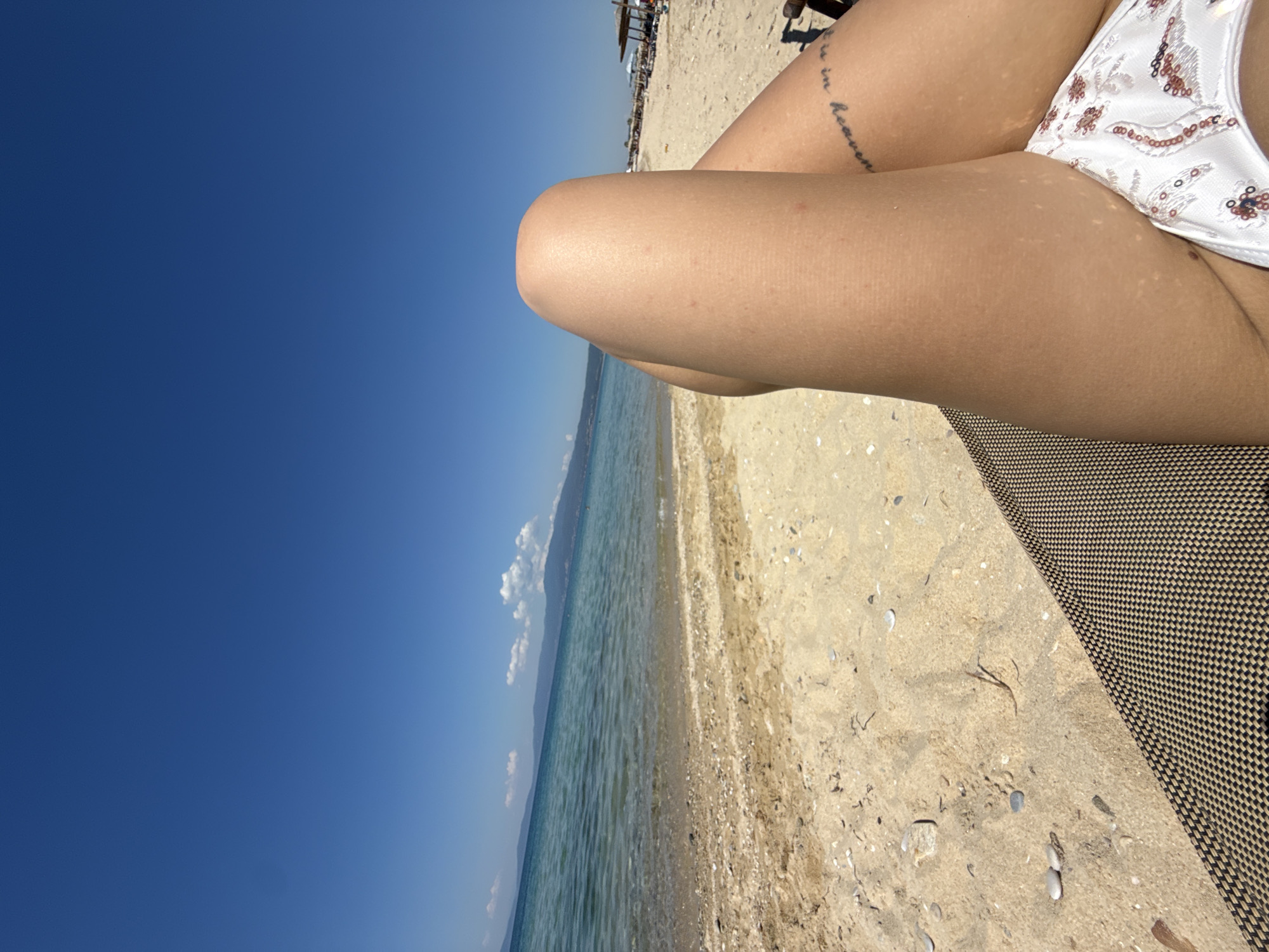 The image shows a beach scene. A person is sitting on a brown woven beach chair, with their legs crossed in the foreground. The person is wearing a white patterned bikini bottom. A tattoo is visible on their leg. The beach has tan sand, and the ocean is light blue with some white waves. The sky is a clear, dark blue. In the far distance, there are some people and beach umbrellas on the sand. The location appears to be a beach with clear skies and calm waters, somewhere in an unknown city and country.