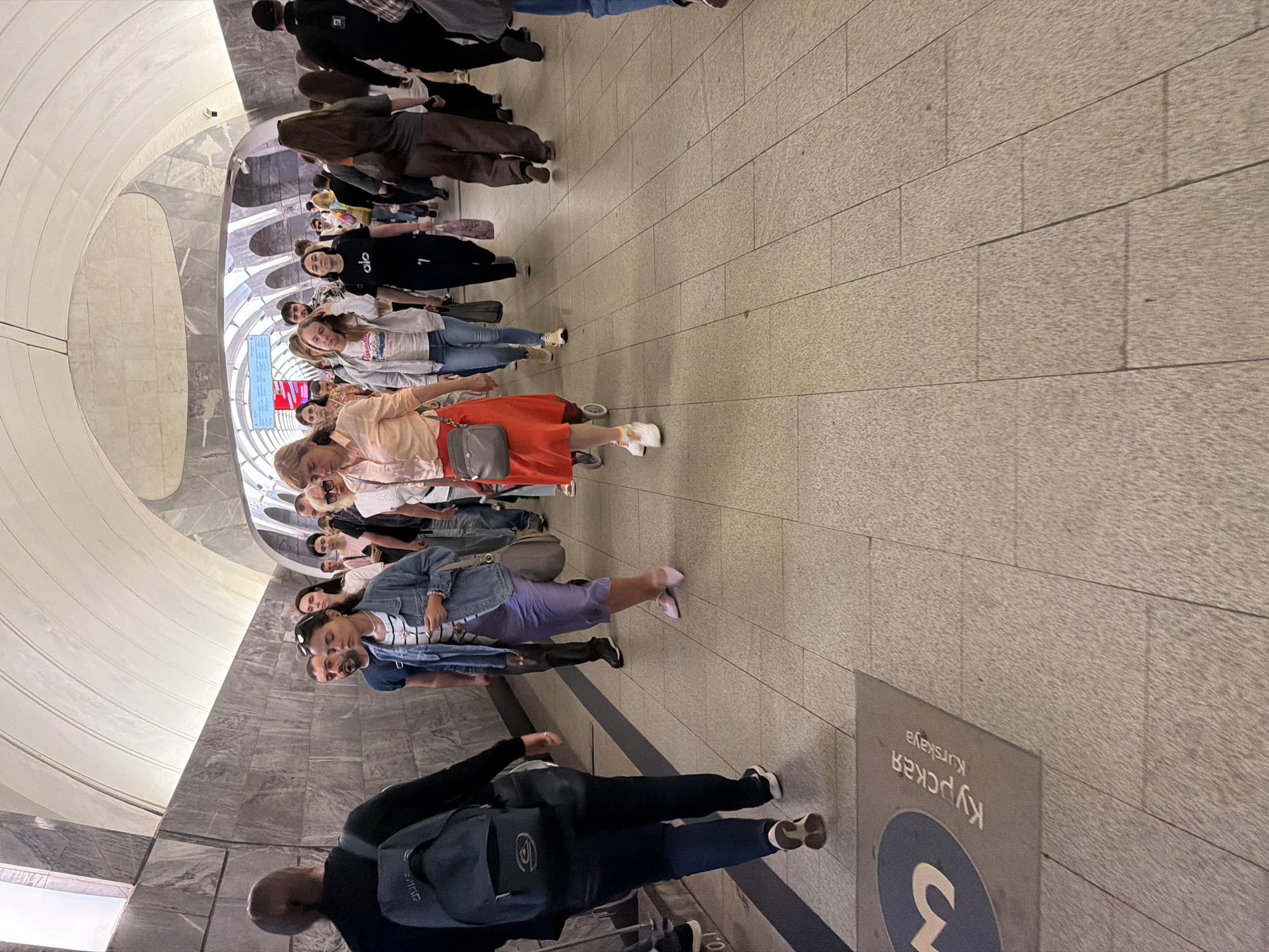 The image depicts a busy underground pedestrian tunnel. The tunnel has a curved ceiling and walls clad in grey marble. The floor is paved with large, light-colored tiles. A diverse crowd of people, dressed in casual clothing, are walking in both directions. Some individuals are carrying backpacks or handbags. In the foreground, a mat on the floor features the number '3' and text in Russian and English that reads "Kurskaya" and "Kurskaya" respectively, indicating the location as Kurskaya metro station in Moscow, Russia. The lighting appears to be artificial, illuminating the tunnel and the people within it.