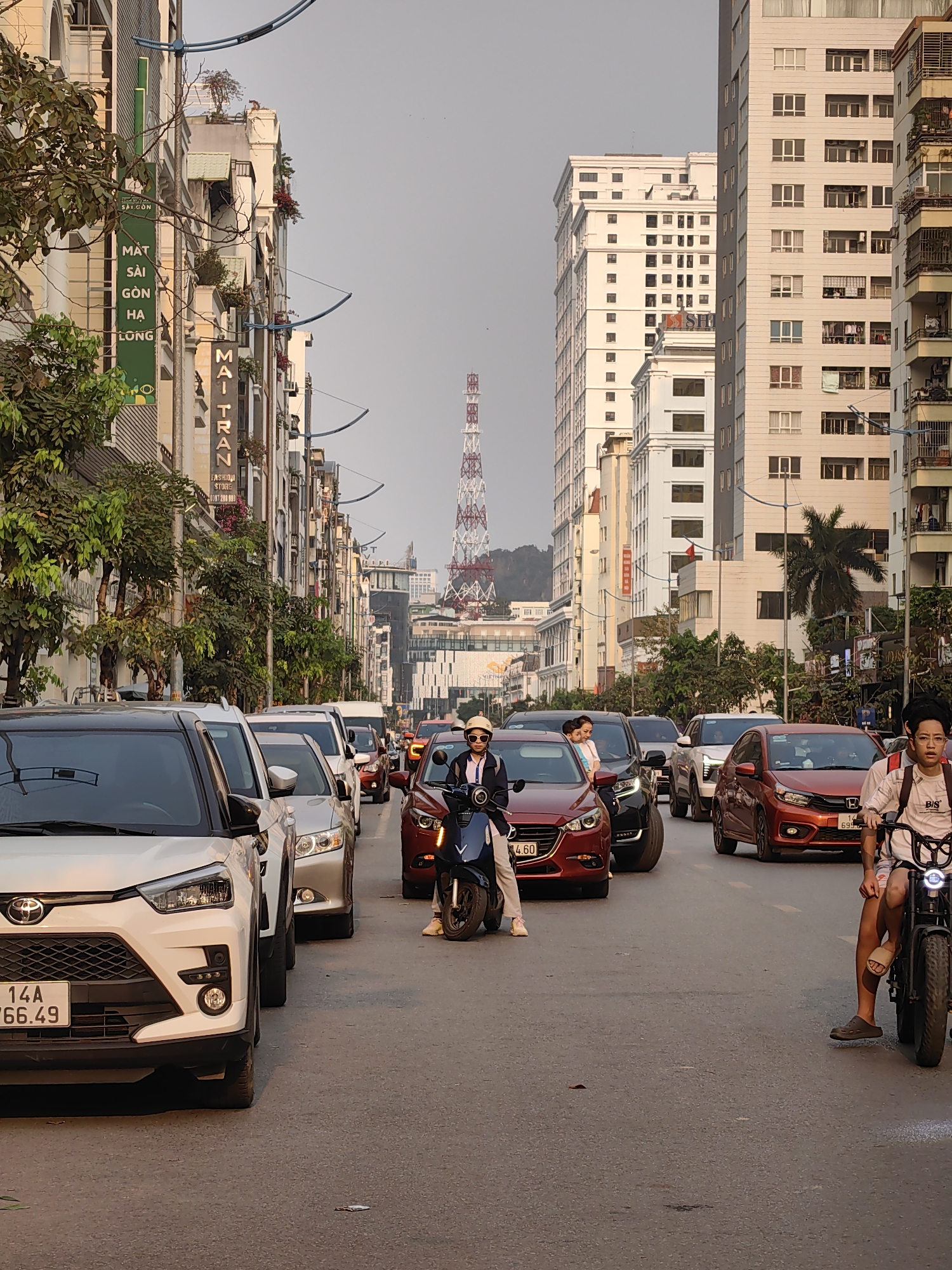 The image captures a street scene in Hong Gai Ward, Vietnam, characterized by heavy traffic. A mix of cars and motorbikes fill the road, with a white Toyota SUV prominently positioned at the front on the left. A woman in a white helmet rides a blue motorbike in the center of the road, and a young man on an electric motorbike is visible on the right. The architecture lining the street includes tall buildings, some with balconies and apartment-style facades. Signs in Vietnamese are affixed to buildings on the left, including one with the words "MAT SAI GON HA LONG." In the distance, a tall, red and white communication tower is visible, set against a backdrop of hills. The sky appears overcast. The license plate on the Toyota reads "14A 66.49." A red car has the license plate "14 H 699.6".