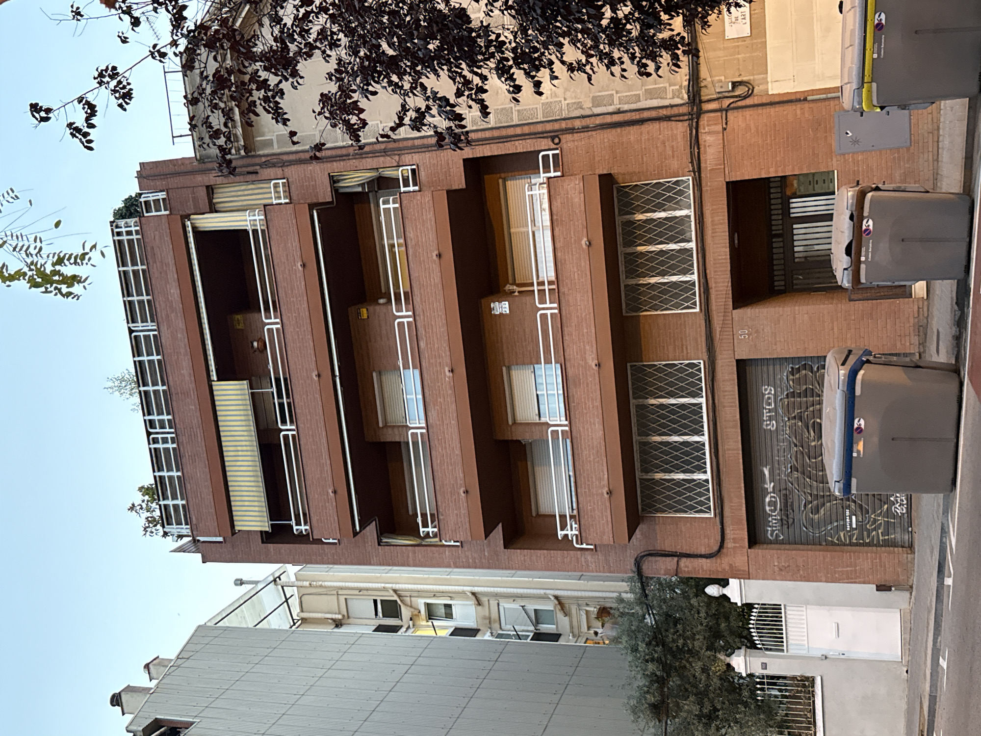 The image shows a multi-story brick building with a stepped facade, likely in an urban setting. The building has several balconies with white railings and some with striped awnings. The windows on the upper floors are white-framed and some have decorative metal grates. On the ground floor, there is a large roll-up door covered in graffiti. Two large grey trash bins are placed on the sidewalk next to the building.
A street sign with text is visible on the upper left side of the building, and the number "50" is painted on the facade near the ground floor entrance. A tree with dark leaves partially obscures the top of the building on the left. Another building with a corrugated metal roof is visible in the foreground on the left, and parts of other buildings and a street are visible on the right. The sky is a pale blue, suggesting it is daytime, possibly late afternoon or early morning due to the lighting. The overall scene appears to be a residential or mixed-use urban environment. There are no people visible in the image. The graffiti on the roll-up door is the only significant text visible, along with the street sign and the building number.