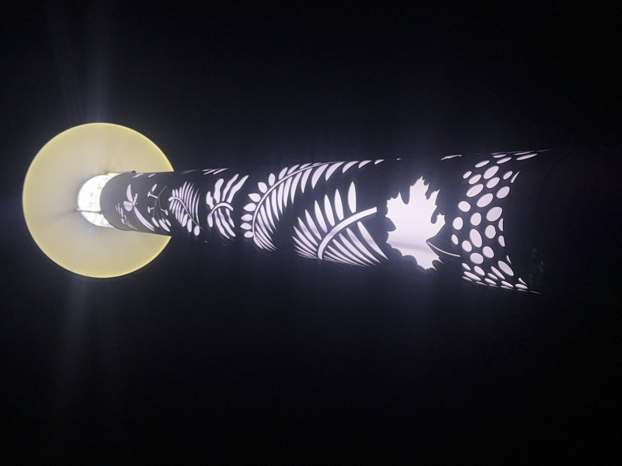 The image depicts a cylindrical lamp in Yekaterinburg, Russia. The lamp has a black exterior with white leaf-like patterns, including maple leaves and ferns. Light shines through the cut-out patterns. A yellow disc is located at one end of the lamp. The background is dark.