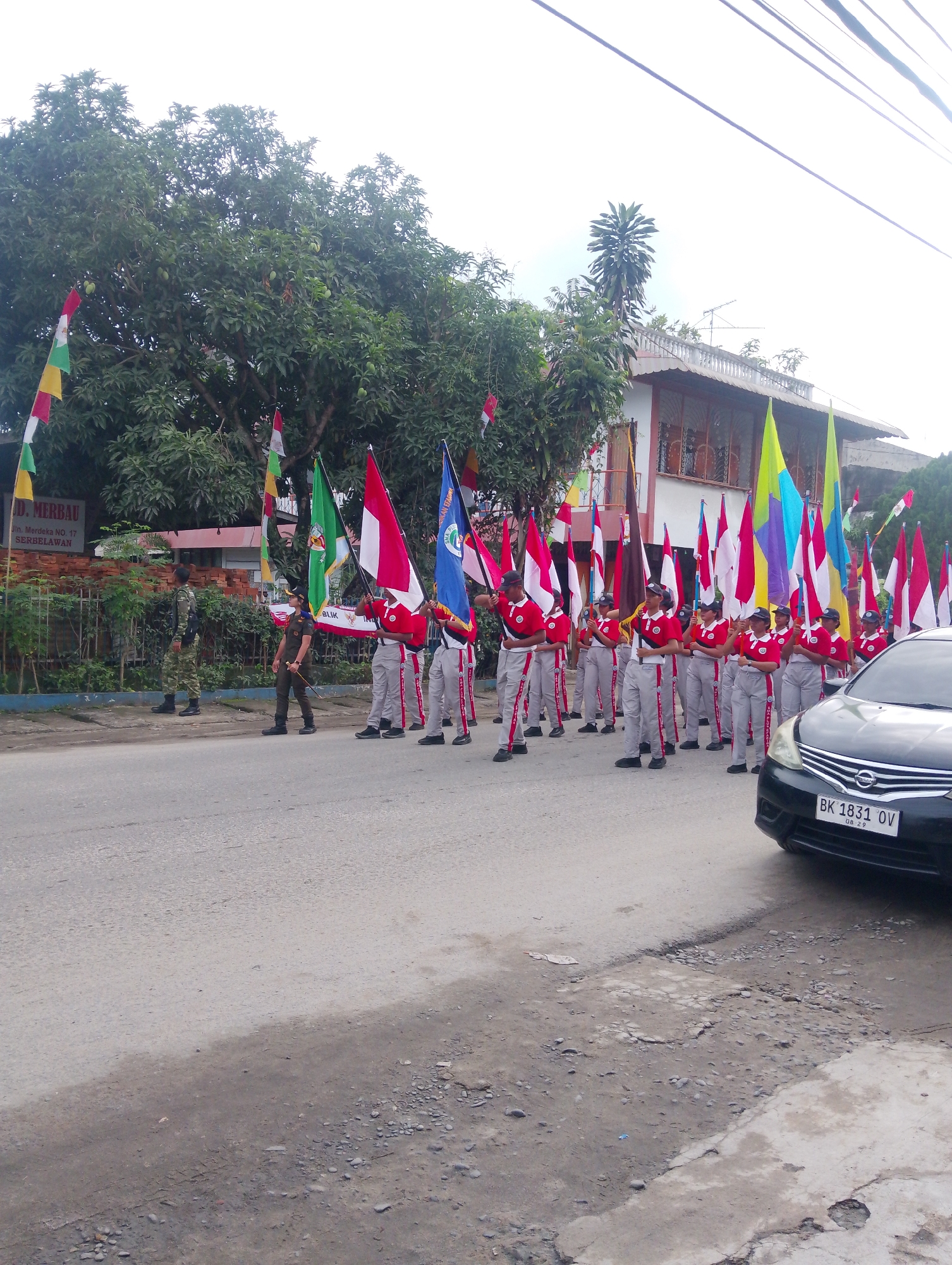 A contingent of marchers, predominantly young people in uniform, is parading along a street. The marchers, some carrying flags and others holding batons, are positioned on the asphalt road, moving from left to right in the frame. Their uniforms consist of red tops with white collars and grey trousers adorned with red stripes. A few individuals in military-style uniforms are also present, observing the procession. The backdrop includes a building with a tiled roof, trees with dense foliage, and various national flags and banners displayed along the street and on poles. A black car with the license plate "BK 1831 OV" is parked on the right side of the road, partially obstructing the view of the marchers. The scene appears to be taking place in an urban or semi-urban environment in Serbelawan, Indonesia, as indicated by a sign visible on the left which reads "JL. MERBAU NO. 17 SERBELAWAN". The sky is overcast.