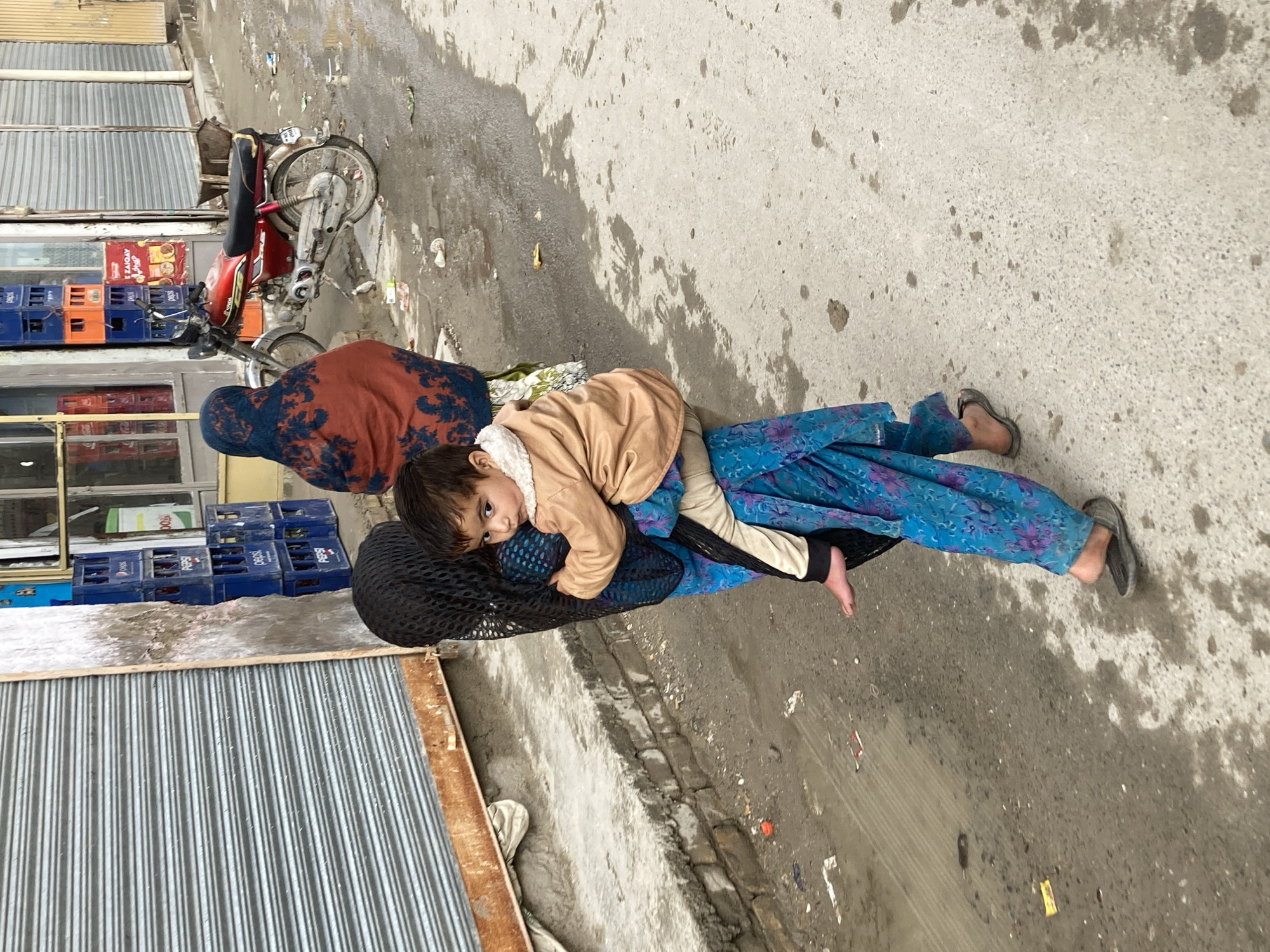 A child wearing a tan jacket is carried on the back of an adult wearing blue patterned pants and a black mesh head covering. The adult is walking on a street in Quetta City Tehsil, Pakistan, in front of a building with a corrugated metal shutter. A red moped is parked nearby. There are Pepsi crates stacked near the building. The ground is wet and littered with debris.
