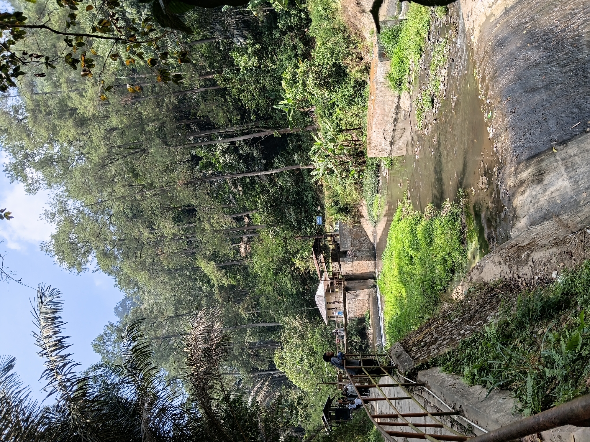 The image shows a view of a dam and a river in the city of Ciburial, Indonesia. The dam is made of concrete and has a stone embankment. A walkway with a railing runs along the left side of the image, with people walking on it. A small building with a white roof is visible behind the dam, and there are trees and foliage on the surrounding hills. The river appears to be flowing and has some debris in it. The sky is visible at the top of the image, with some clouds.