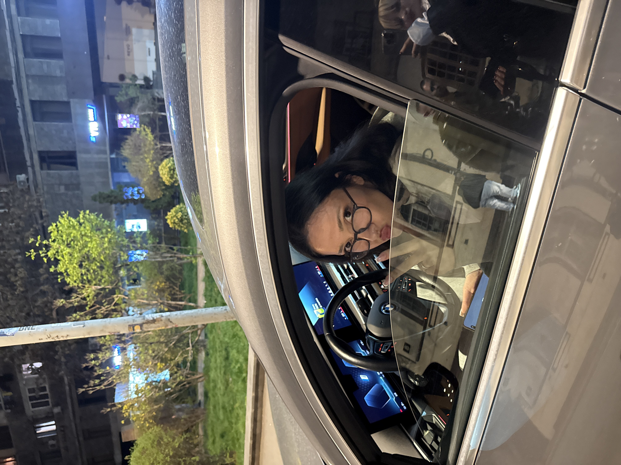 A woman with glasses is sitting in the driver's seat of a gray car. She is looking directly at the camera. The car has a digital display and a BMW steering wheel. The background shows a cityscape at night with buildings and trees lit by streetlights. Street signs display "ONE" and the Yerevan city name in Armenian. Based on the surroundings, the location is Yerevan, Armenia.