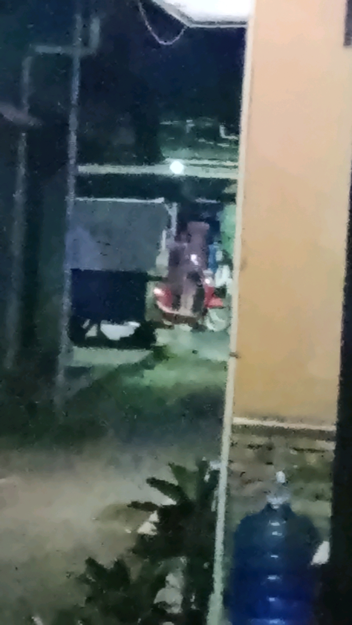 The image, taken at night in Ujungrusi, Indonesia, shows a blurry scene with a focus on a red scooter with people on it. A large, dark rectangular object, possibly a truck or a cart, is positioned behind the scooter. To the right of the scooter, a tall, light-colored structure, likely part of a building, is visible. In the foreground on the far right, a large blue water dispenser is partially visible. The ground appears to be paved and dimly lit, with some sparse vegetation in the lower left quadrant of the image. The overall lighting is low, suggesting it is nighttime.