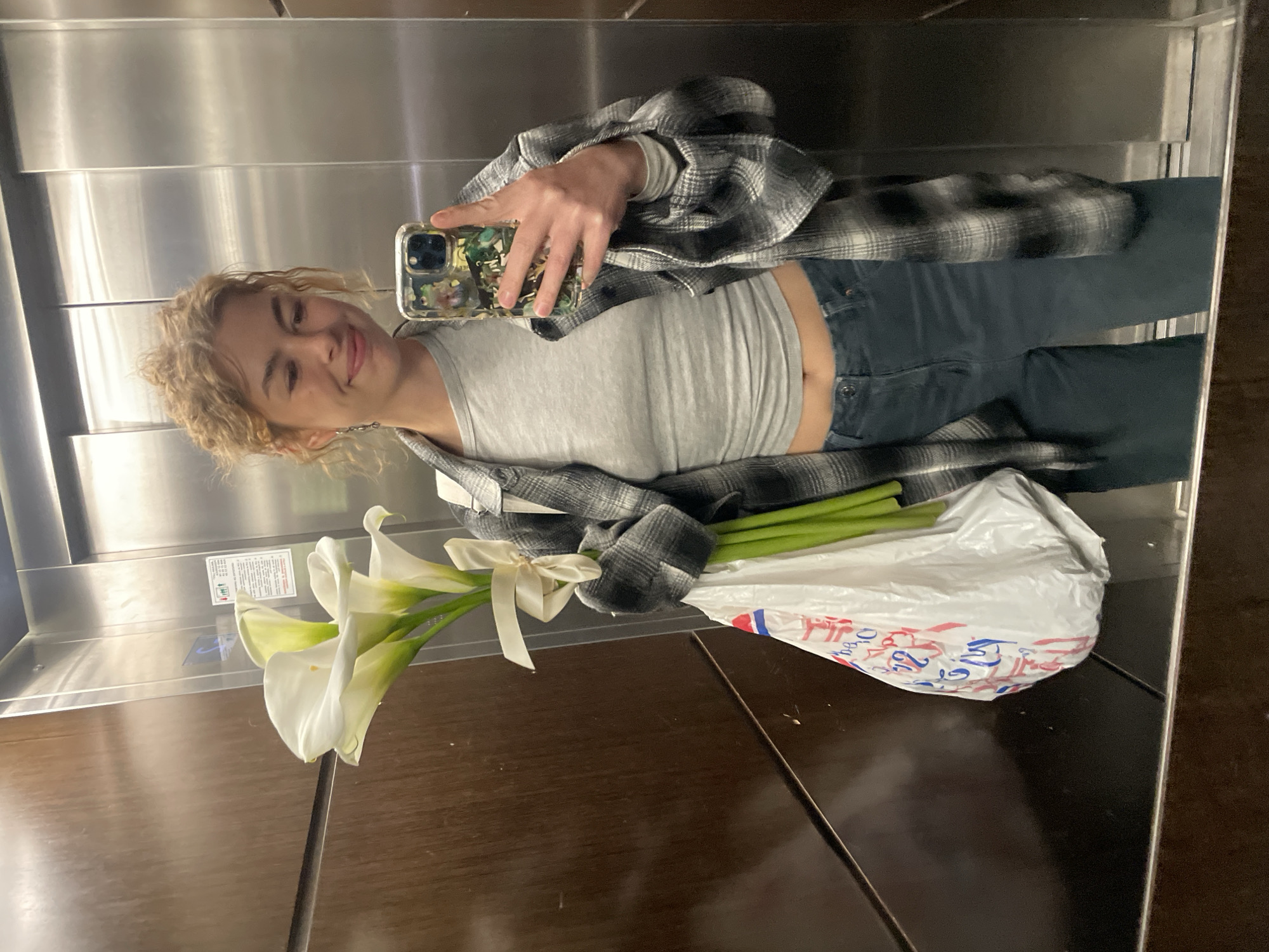 The image captures a mirror selfie taken by a young woman in an elevator, rotated 90 degrees clockwise. The woman, with light curly hair, is looking into the camera with a gentle smile. She is wearing a grey crop top, a black and white plaid button-up shirt, and dark pants. Her right hand holds a smartphone with a distinctive, patterned case, used to take the picture.
In her left arm, she holds a bouquet of several elegant white calla lilies, neatly bound with a cream-colored ribbon. Next to the flowers, she carries a white plastic bag featuring partially visible red and blue lettering that appears to read "Тропи". The background reveals the polished stainless steel walls of the elevator, which serve as the mirror, and a dark wooden or laminate floor. An elevator information panel with some unreadable text, arrows, and a blue symbol is visible on the wall. The overall activity is a casual mirror selfie, possibly taken while traveling in an elevator, perhaps after purchasing flowers or returning from an event. The woman's expression is pleasant and relaxed. The location is specified as Boreti, Montenegro.