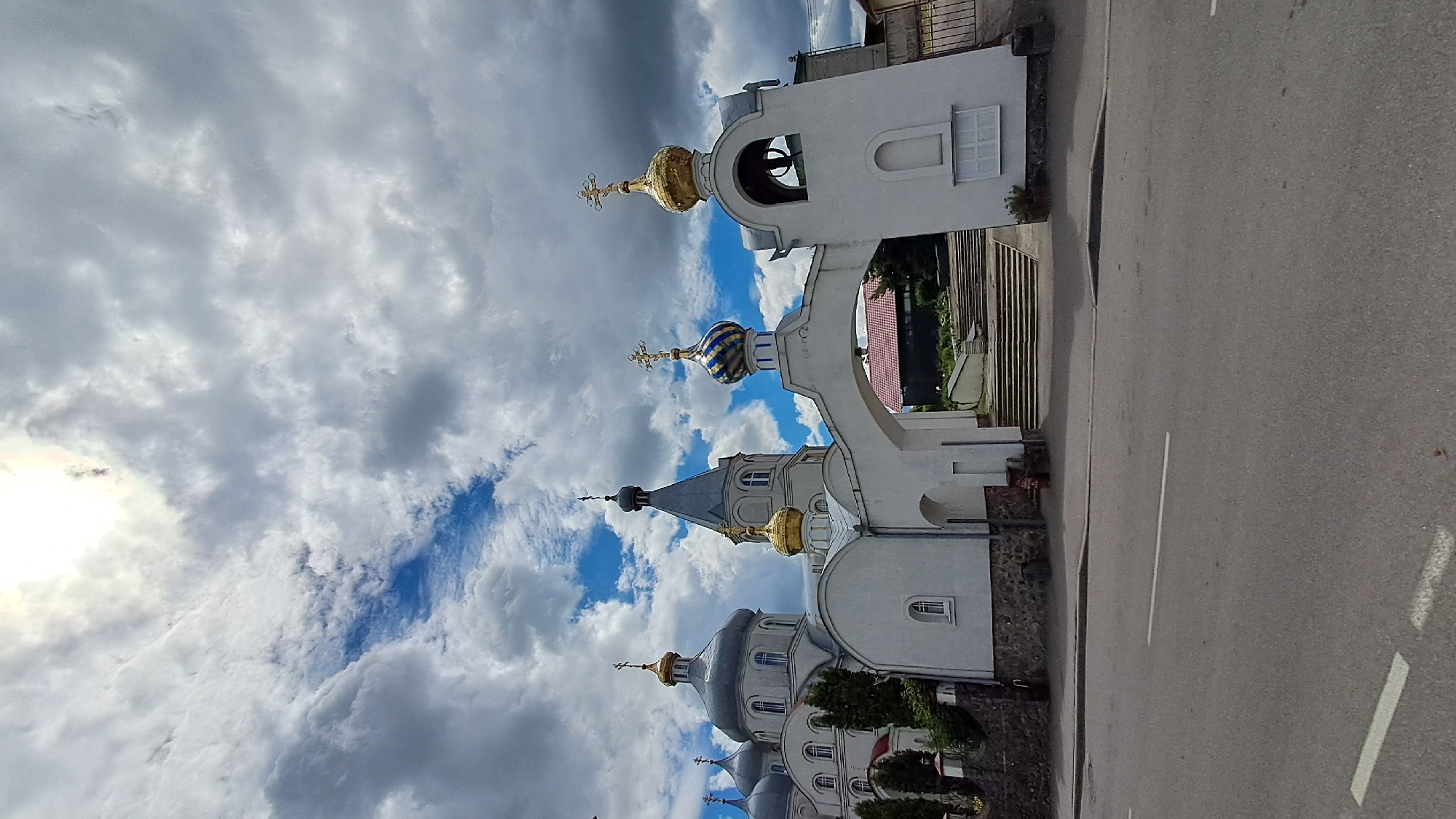 This media file depicts a view of an Orthodox church, specifically the Church of the Assumption of the Blessed Virgin Mary in Vinnytsia, Ukraine. The church features multiple domes, some of which are gilded and others are blue and gold striped. A white bell tower with an arched opening is also visible. The building appears to be constructed of light-colored stone or stucco.
The scene is set outdoors, with an asphalt road in the foreground. The road has white markings. Behind the church, there are some trees and a hint of other buildings. The sky is dramatic, filled with a mix of dark and white clouds, with patches of bright blue sky peeking through. The sun is partially obscured by clouds, suggesting it might be late afternoon or early evening, or a day with mixed weather. The overall impression is one of a solemn and perhaps slightly overcast day. There is no visible text in the image.